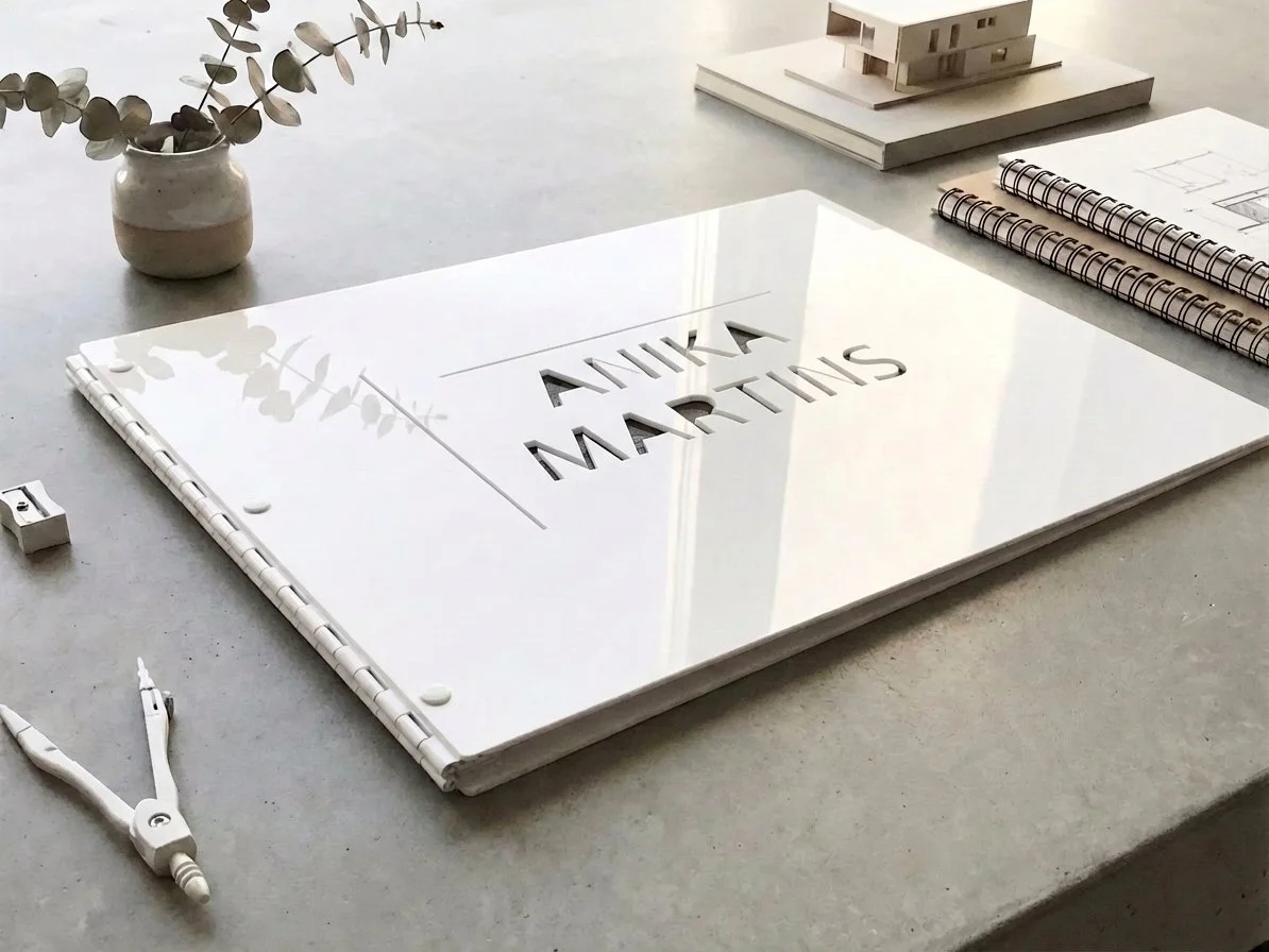 A white presentation board with cut-out letters spelling 'AYUKA MARTINS' on a desk. There are architectural sketches and a small model of a house, a vase with eucalyptus branches, a pair of compasses, and some spiral notebooks nearby.