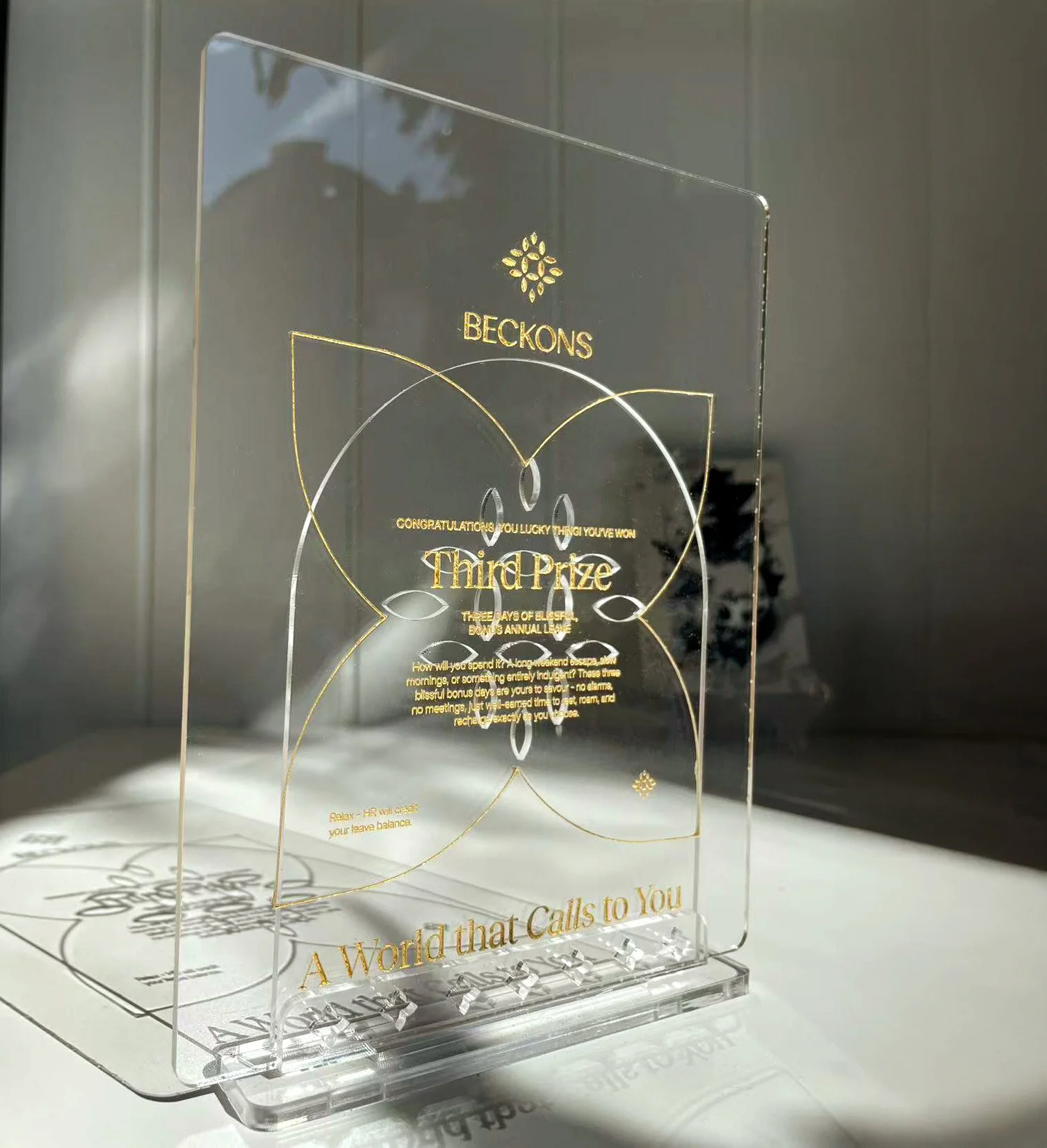 Clear acrylic award display with gold printed details and elegant engraved linework, presented on a transparent stand.
