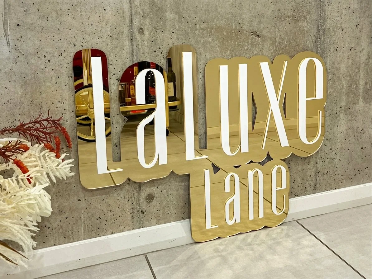 Decorative sign that reads 'Luxe Lane' mounted on a concrete wall, with a mirror reflected in the sign and a white floral arrangement nearby.