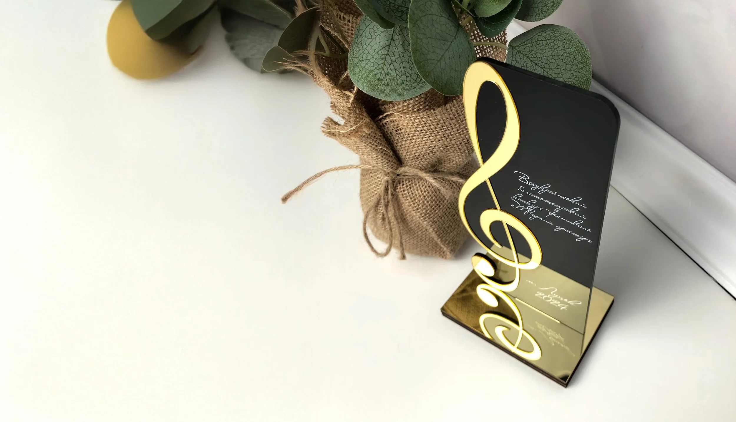 Black and gold trophy with a musical note shape, placed on a reflective base, next to a plant wrapped in burlap and tied with string, on a white surface.