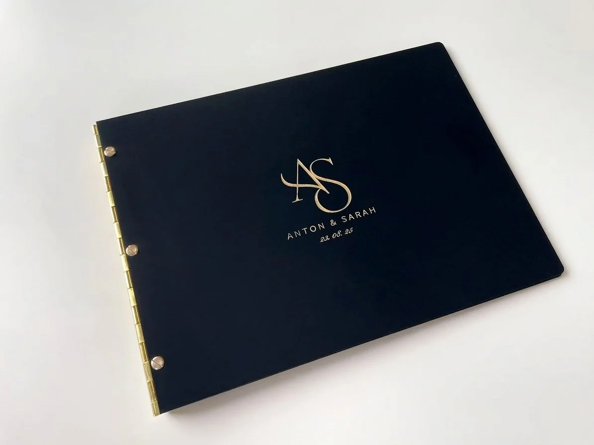 A black photo album with gold binding and personalized engraving on the cover that says 'ANTON & SARAH 23.08.25' with a monogram 'AS' in the center.