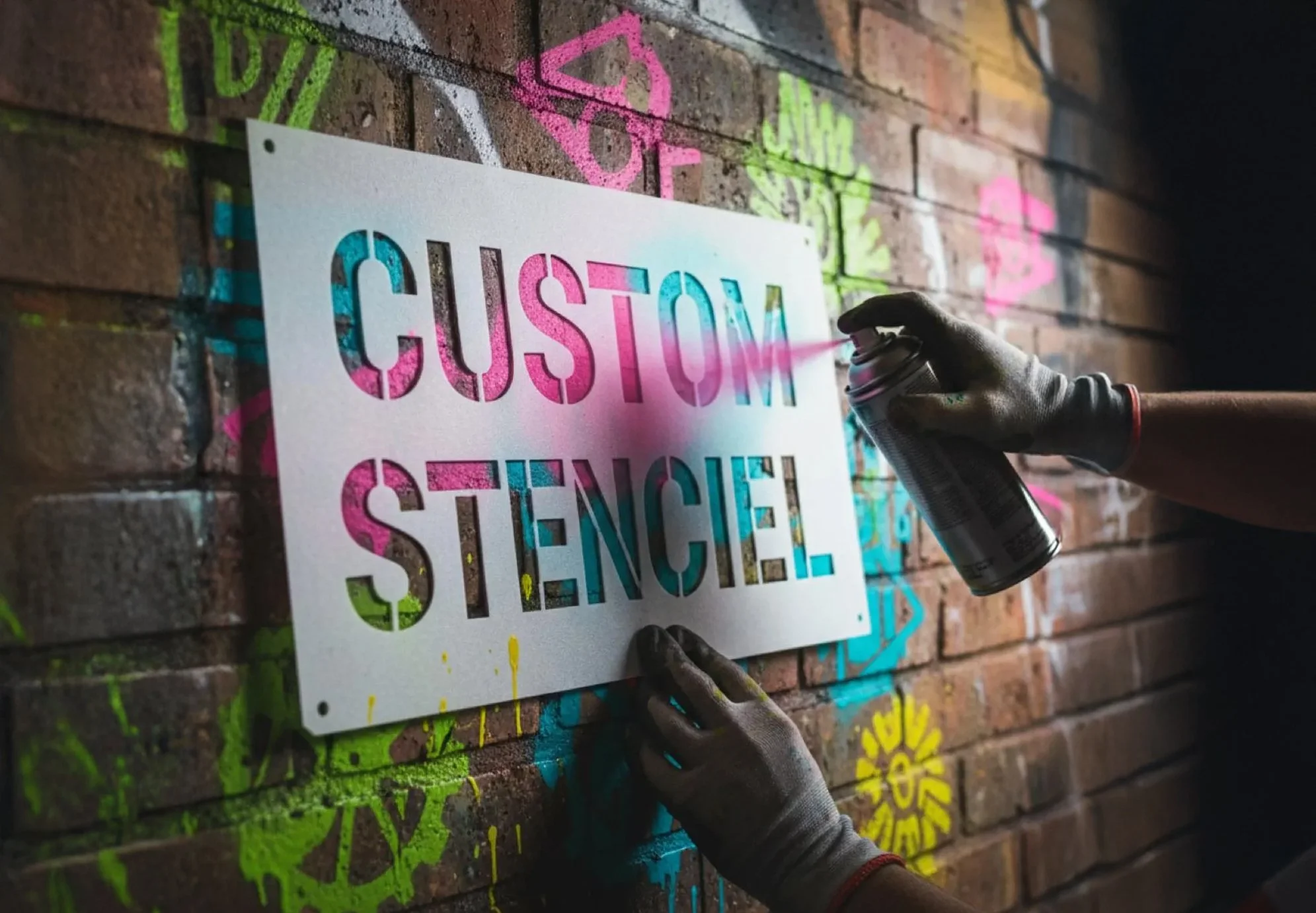 Person spray painting a sign that says 'Custom Stencil' with colorful graffiti on a brick wall.