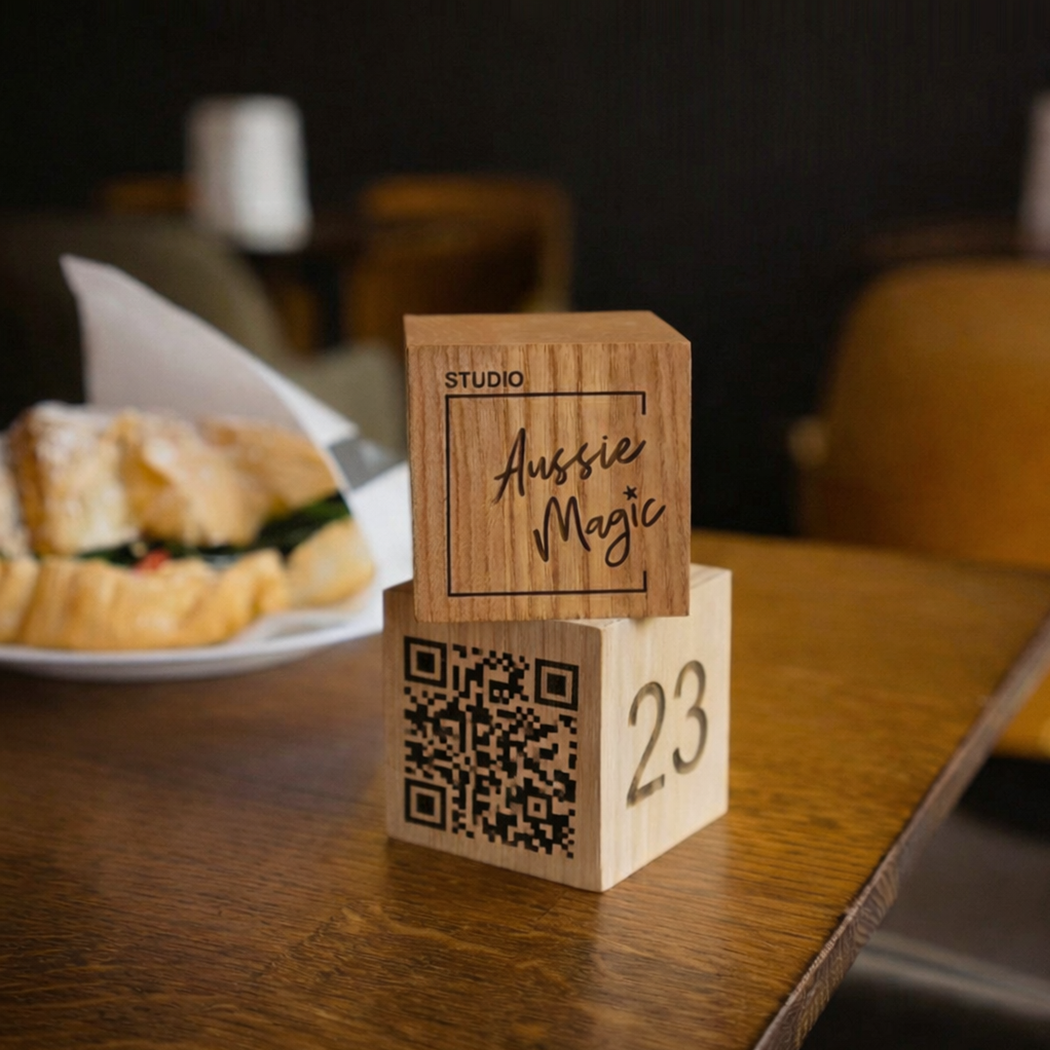 Wooden tabletop display block with engraved QR code, table number and branded top cube shown in a café setting
