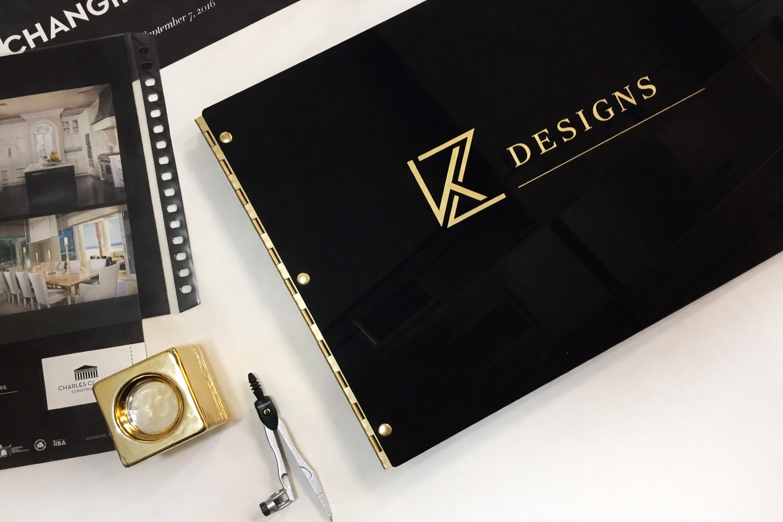 Black presentation board with gold lettering and logo, a magnifying glass with a gold frame, a pair of compasses, and a sheet of photographs of interior design projects on a white surface.