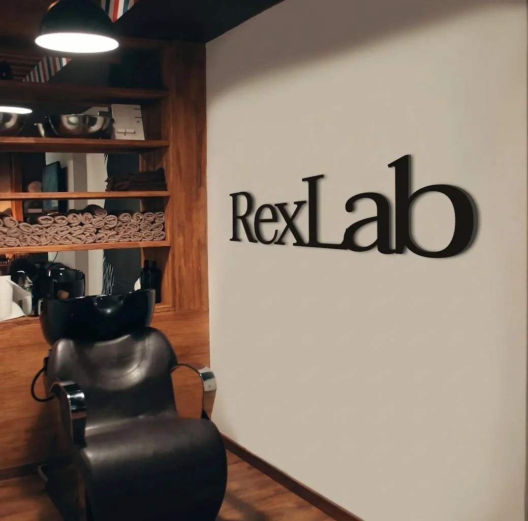 Interior of a salon with a black chair, a wall-mounted sign reading 'RexLab', wooden shelves with towels and salon equipment, and a hair washing bowl.