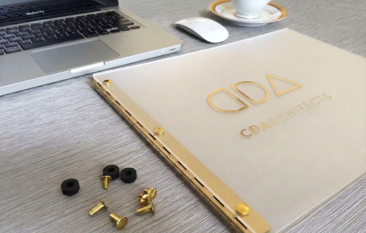 Open CAD architectural plans on a desk with a laptop, mouse, cup and saucer, and small hardware parts including screws, nuts, and washers