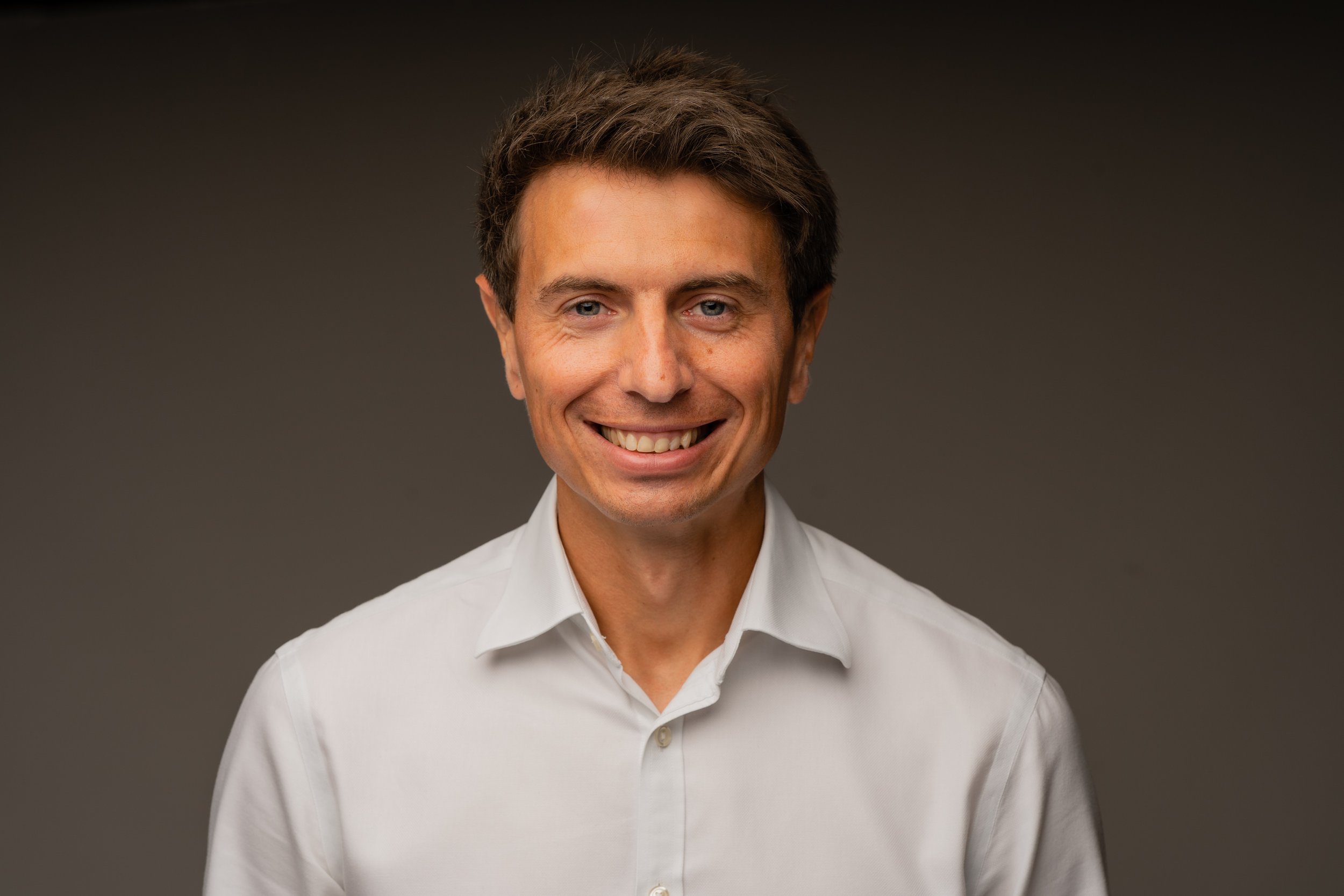 Headshot of a man with brown hair and a white dress shirt smiling against a dark background.
