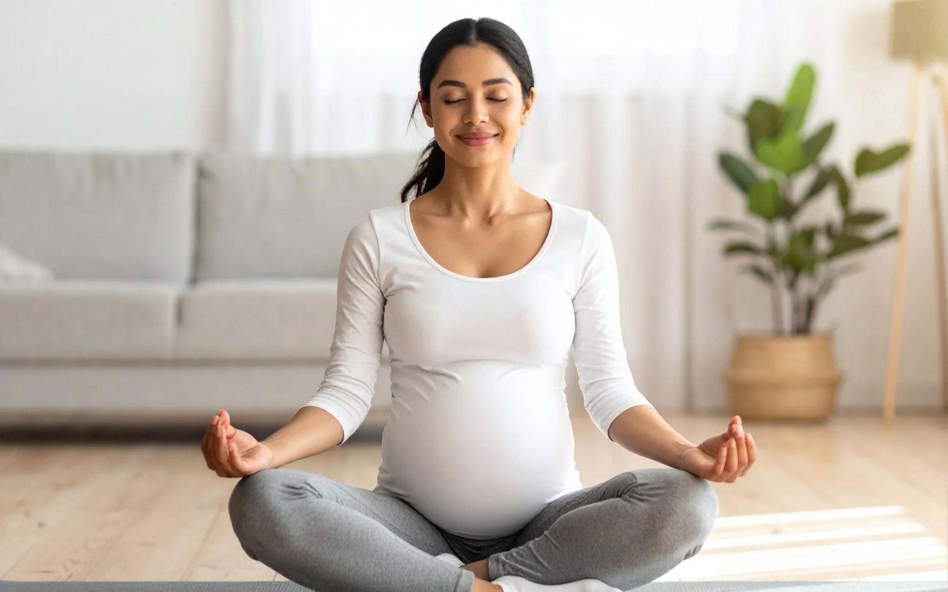Pregnant woman meditating in a cross-legged seated position on a yoga mat in a bright living room.