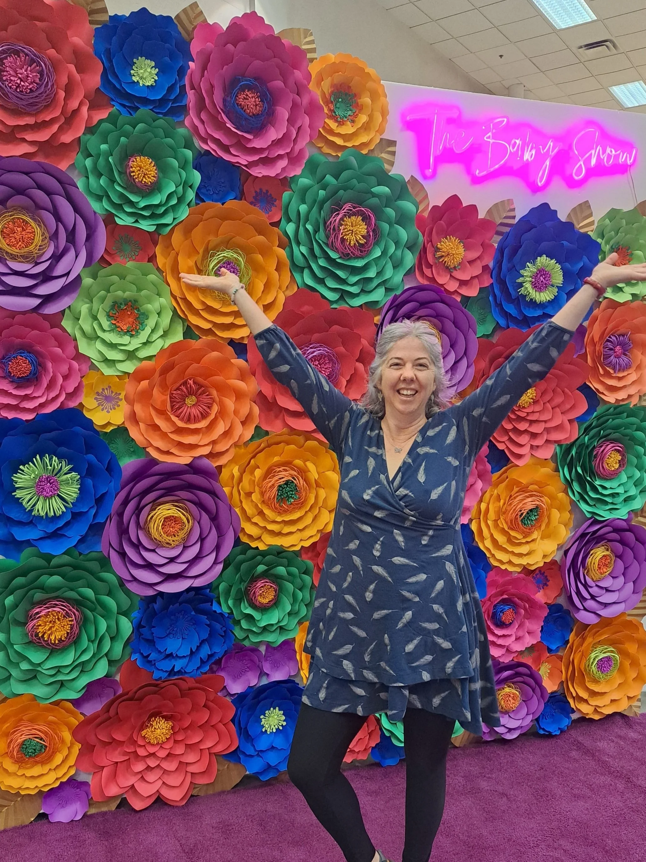 A cheerful woman with gray hair and a dark blue dress with a feather pattern stands with arms raised in front of a colorful paper flower wall at the Toronto Bobbsey Show. The wall features large, vibrant paper flowers in pink, purple, blue, green, red, orange, and yellow hues. A pink neon sign reads 'The Bobbsey Show' in the background.