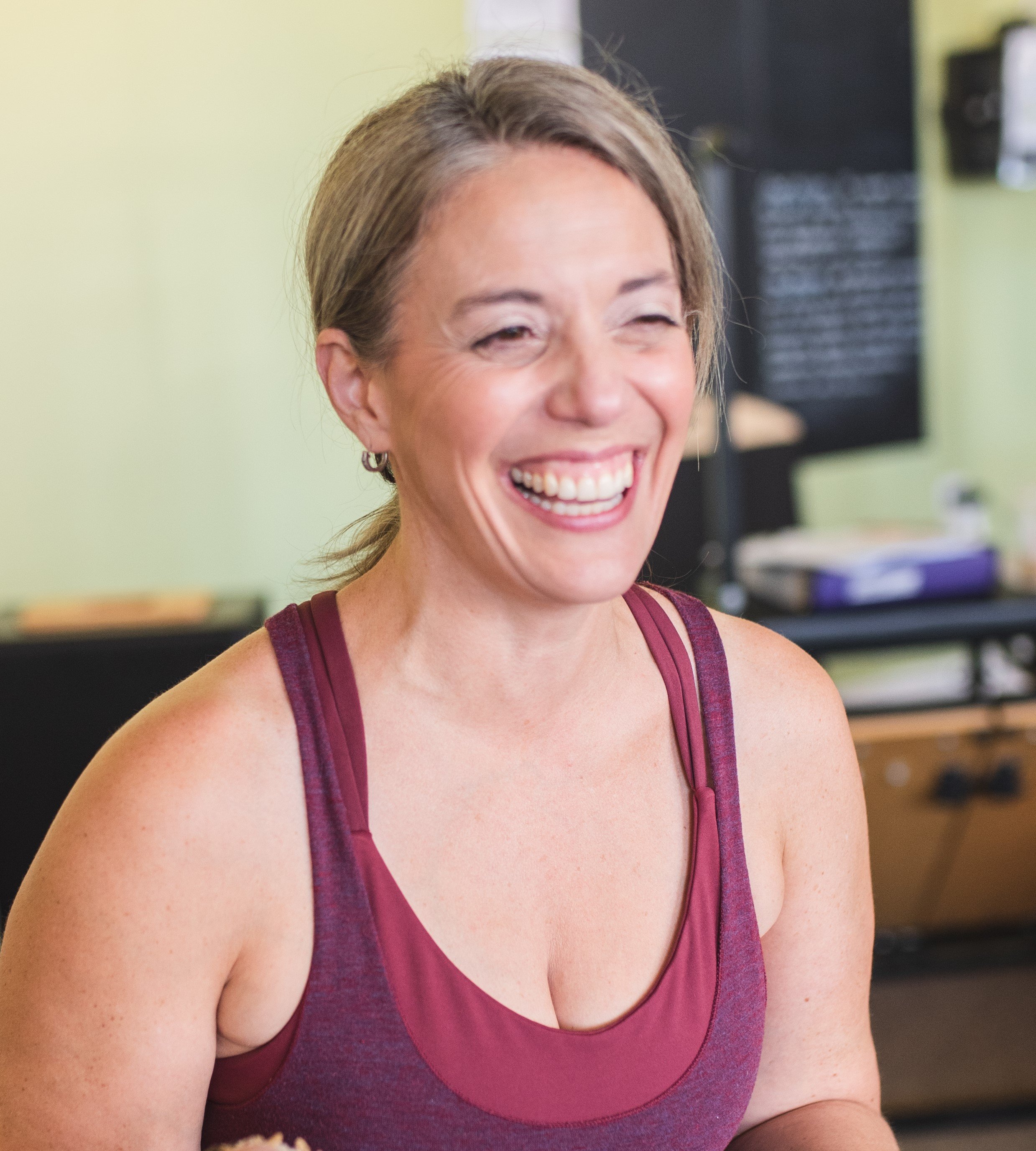 A woman with light brown hair wearing a sleeveless maroon athletic top, smiling and laughing in a gym or sports facility.