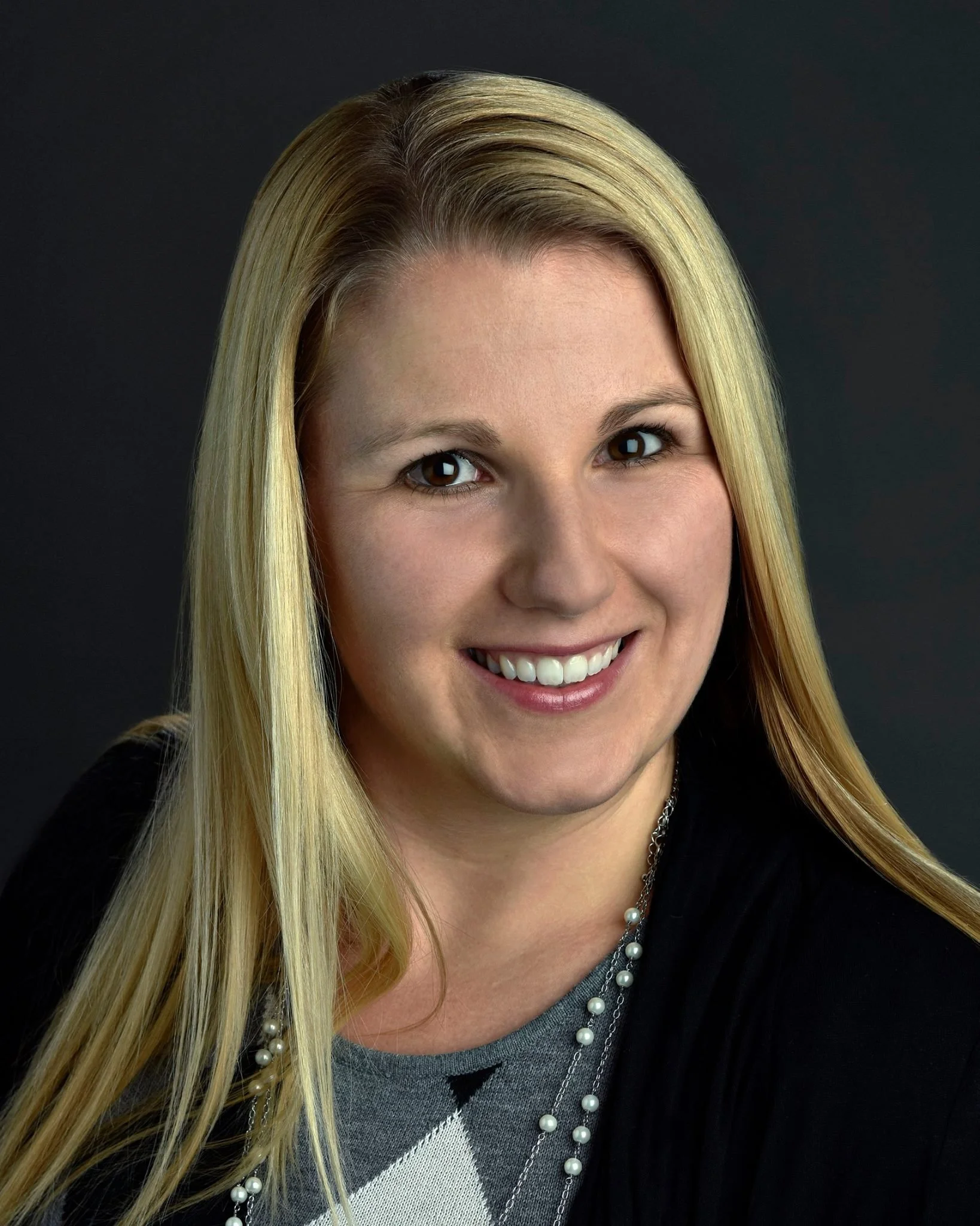 A smiling woman with long blonde hair wearing a black top and pearl necklace against a dark background.