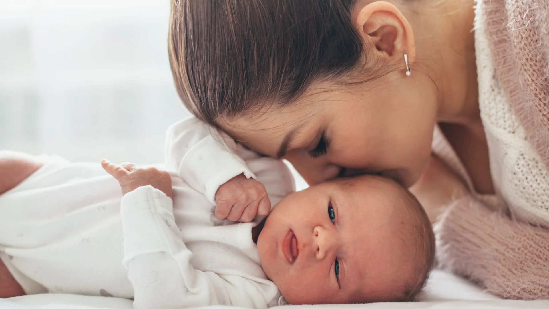 A woman gently kisses a newborn baby on the forehead. The baby is lying on a white surface, wearing a white outfit and looking at the camera. The woman has brown hair and is wearing a pink knitted sweater and small earrings.