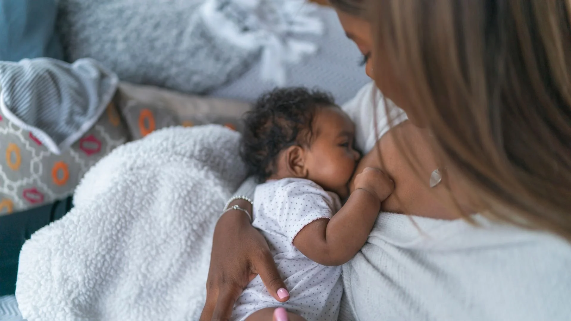 A mother breastfeeds her baby lying on her chest, both lying on a bed with pillows and blankets.