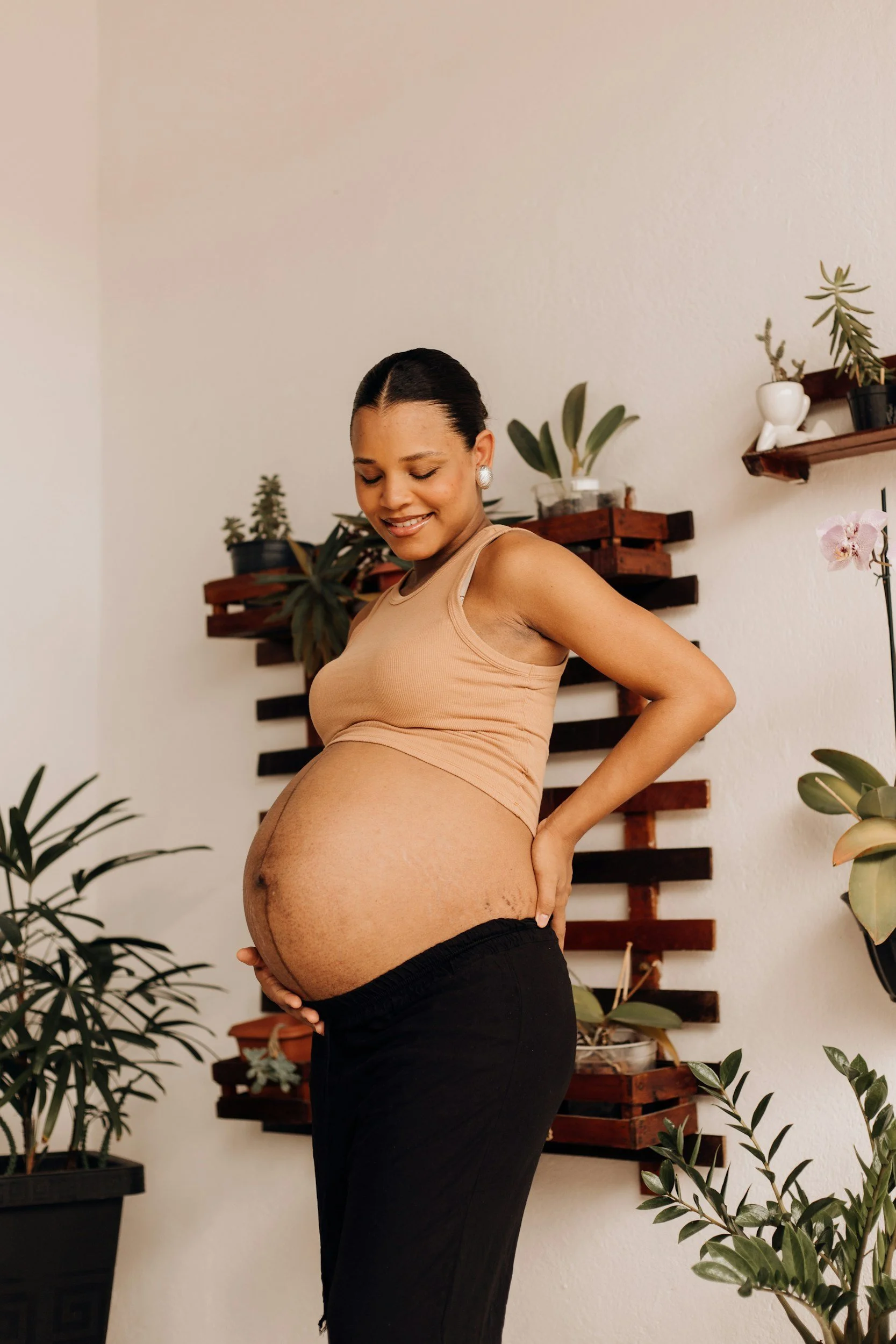 Pregnant woman smiling and holding her belly in a room with plants and wooden wall shelves.
