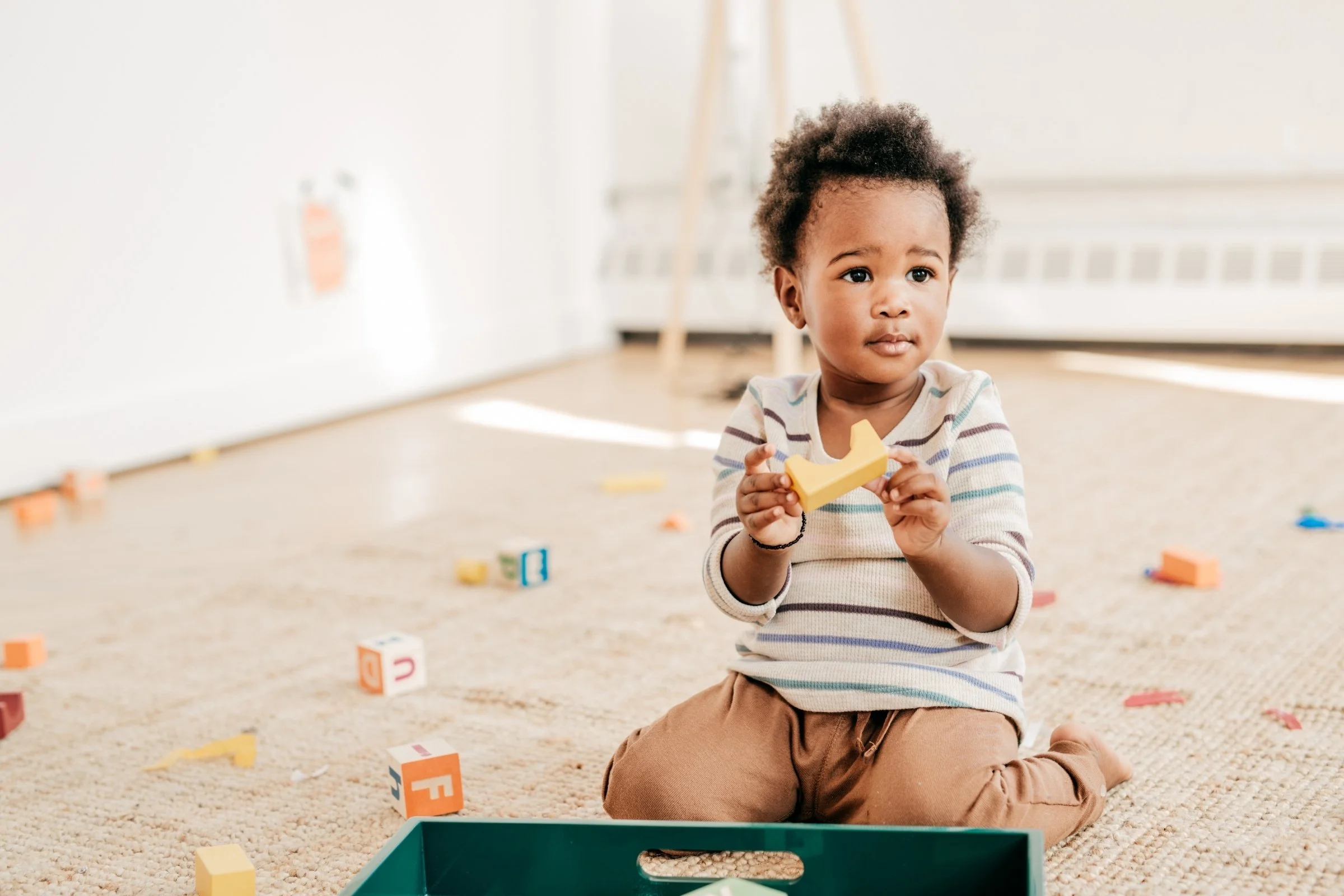 A young child sitting on a carpeted floor, holding a yellow wooden block shaped like a crescent moon, surrounded by scattered colorful alphabet blocks, in a well-lit playroom.