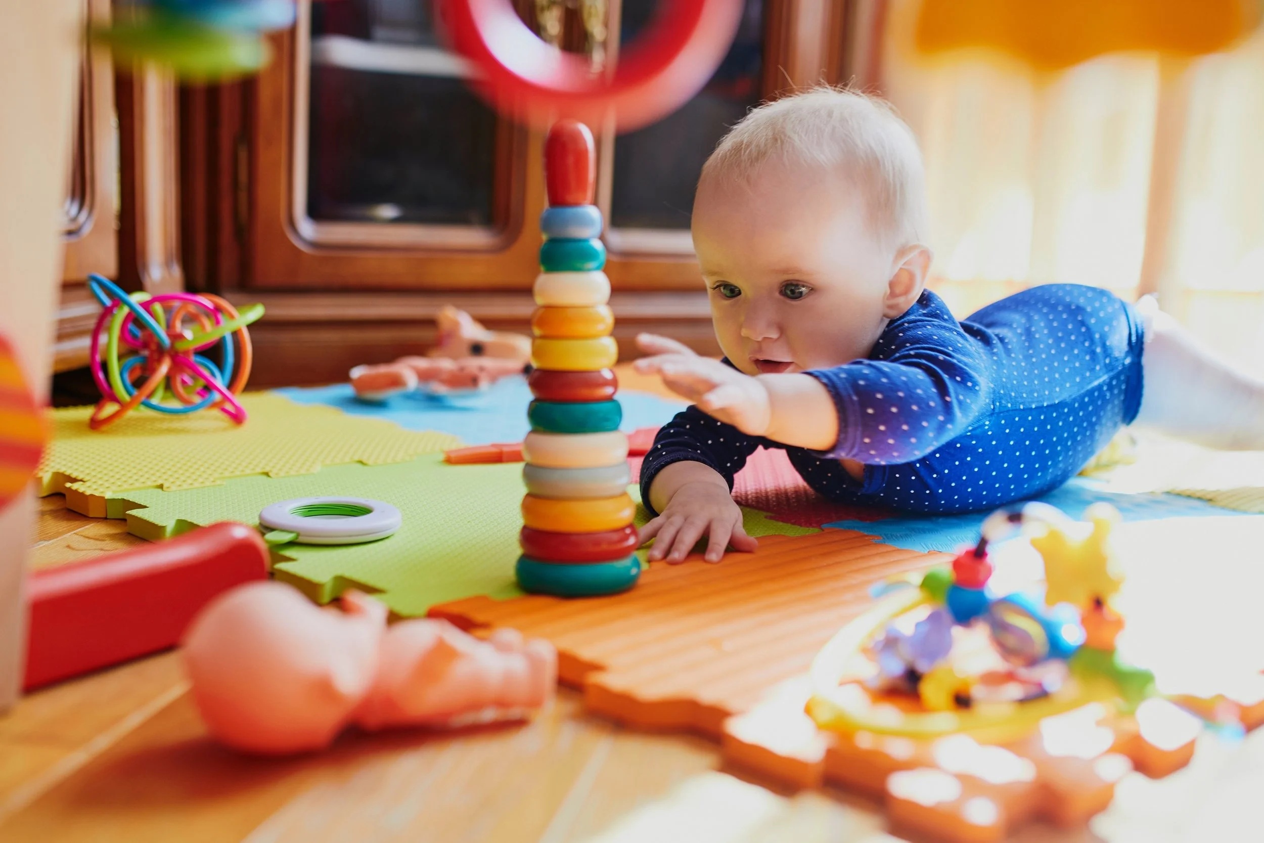 Young child in blue pajamas playing with colorful stacking blocks on a play mat surrounded by toys in a sunny room.