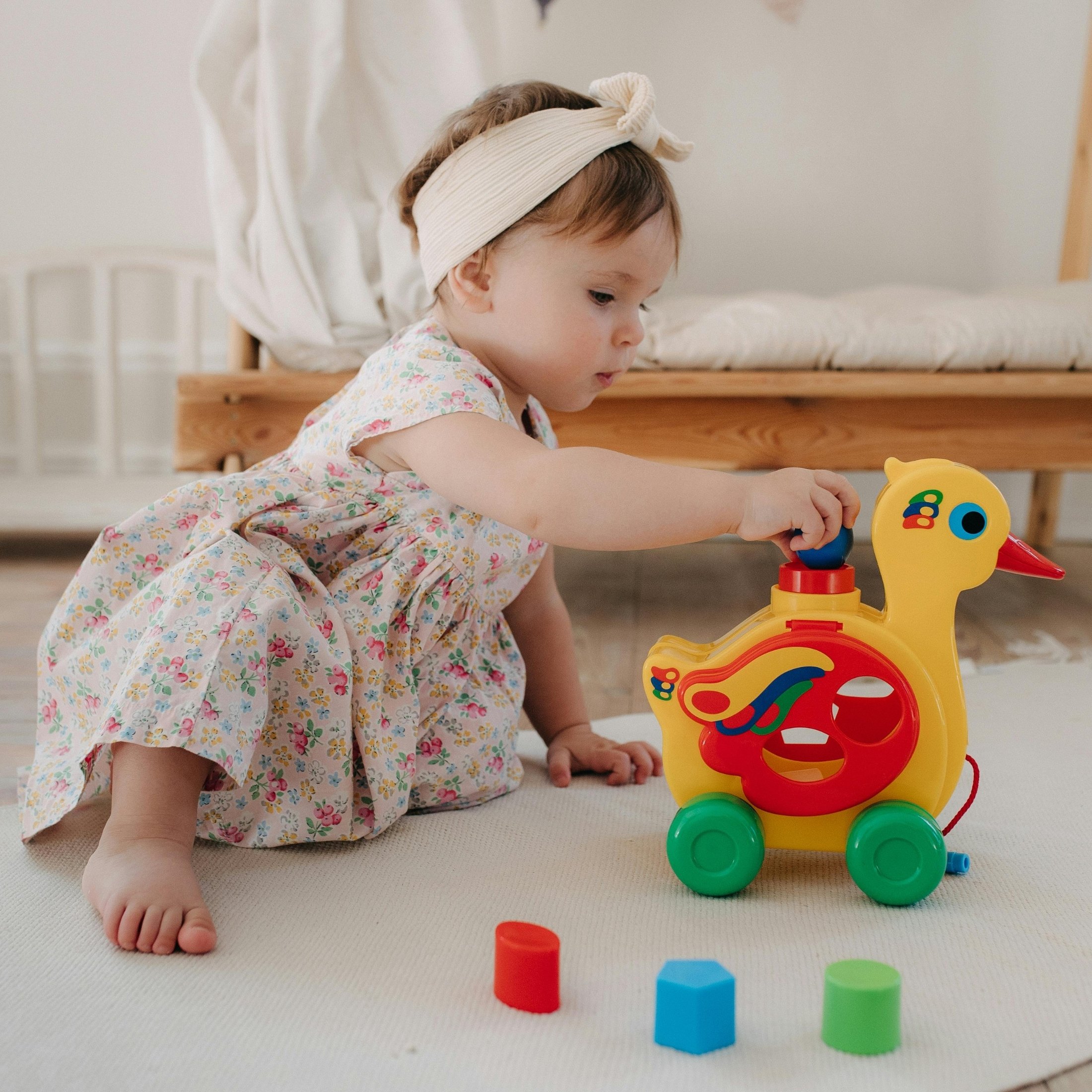 A young girl in a floral dress and headband playing with a yellow toy duck on wheels, inserting one of the colorful blocks into the toy's slot, on a white surface in a cozy room.