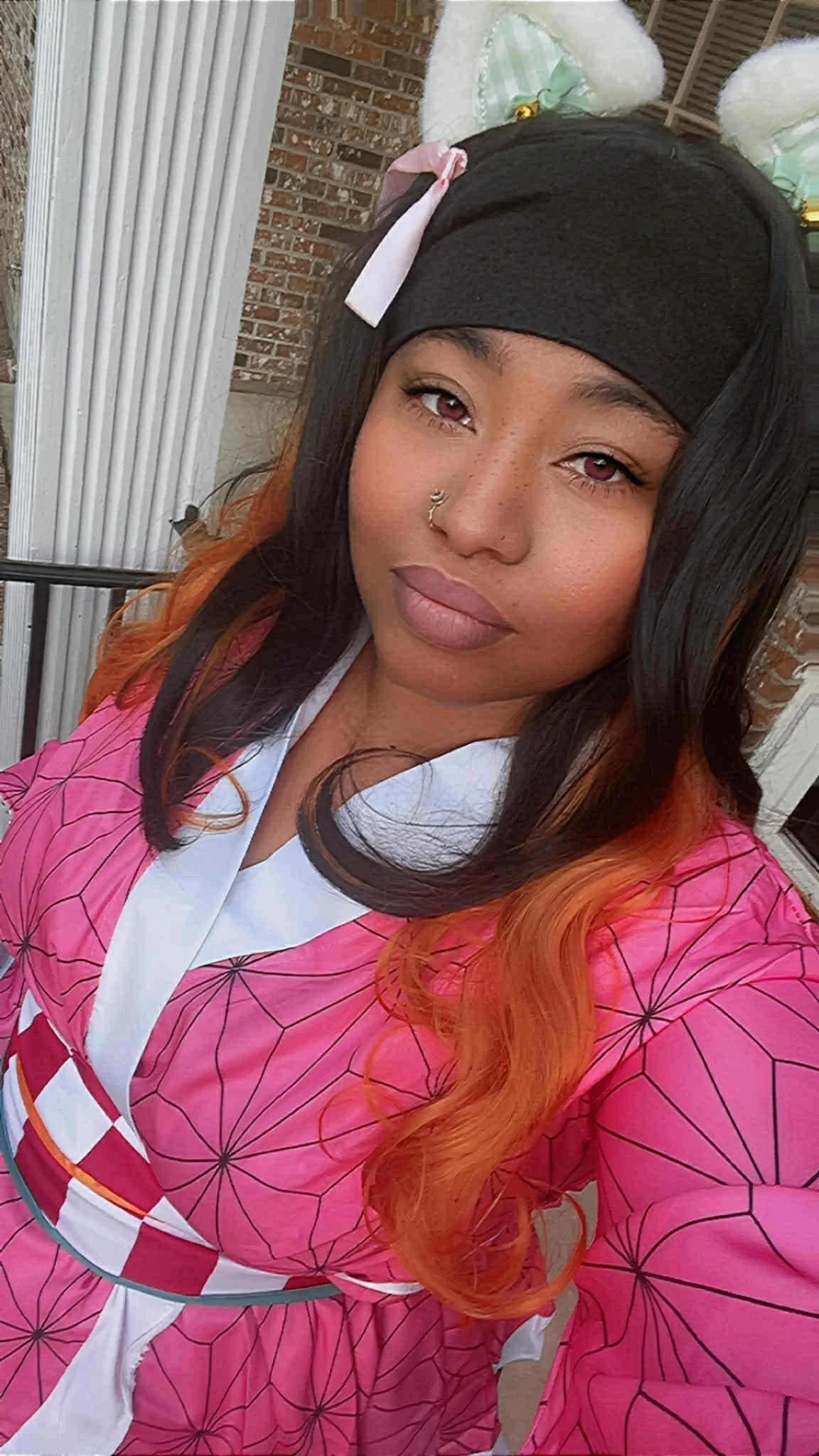 A young woman with pink and black hair, wearing a black headband with white and pink bows, a pink patterned kimono-style garment, and a white collar, standing on a porch with a brick wall and white door frame in the background.