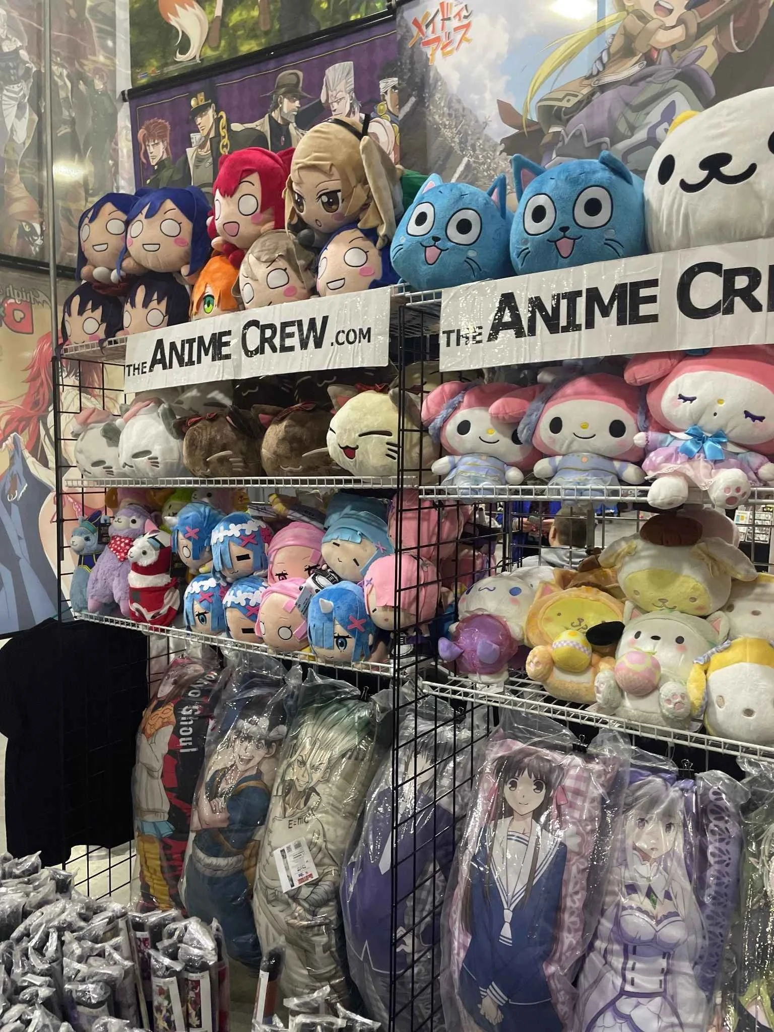 Display of various anime plush toys on a metal rack at a convention, with merchandise bags of anime characters hanging below and anime posters in the background.