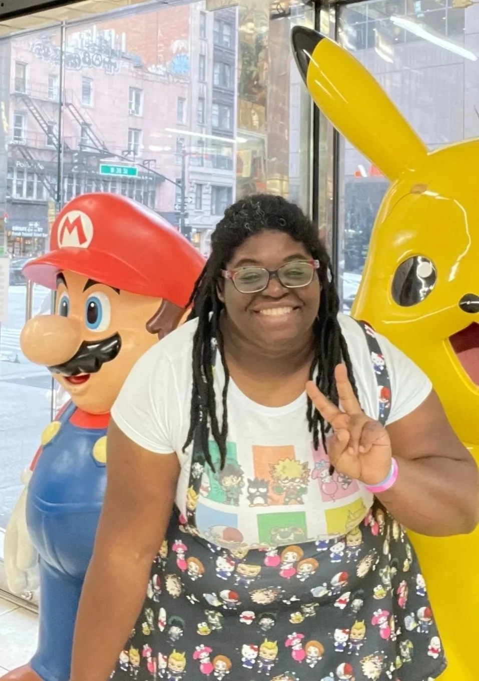 A woman smiling and making a peace sign while posing between a Mario statue and a Pikachu statue inside a store.