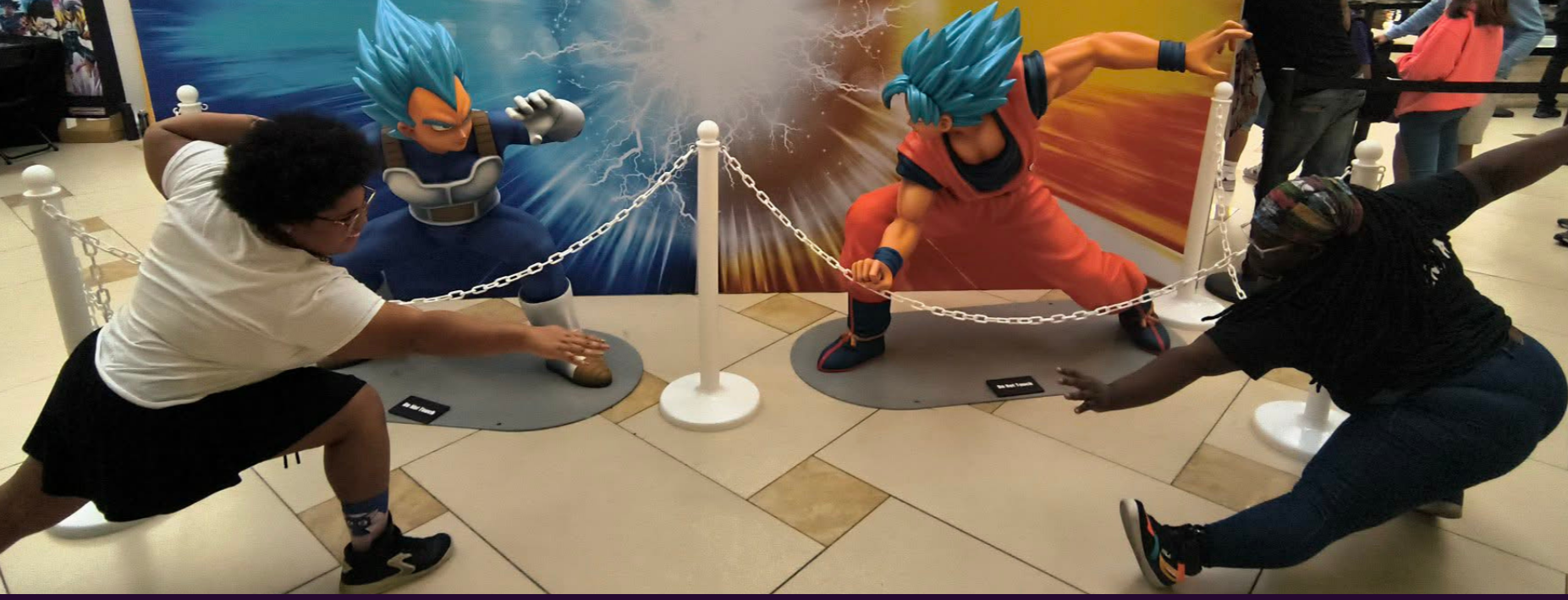 Two people posing in fighting stances with life-sized statue figures of Goku and Vegeta from Dragon Ball. The Goku statue is on the right in orange and blue clothing, and the Vegeta statue is on the left in blue and gray clothing. They are separated by a chain barrier and set against a colorful background.