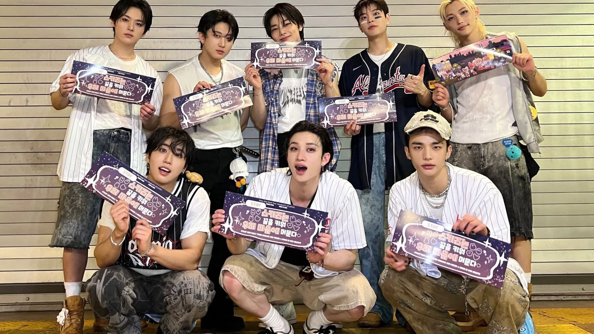 Group of nine young men posing together indoors, holding purple and white signboards with Korean text. They are dressed in casual streetwear, some with blazers, t-shirts, and shorts. The background shows a wall with horizontal lines.