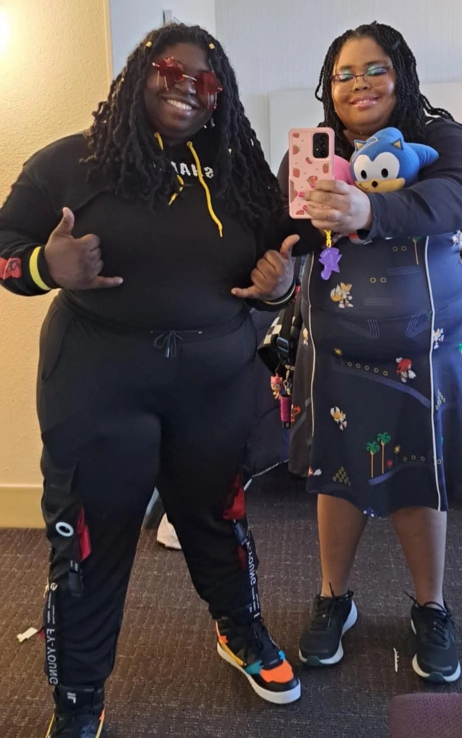 Two women taking a mirror selfie, one in black athletic wear with red sunglasses and the other holding a Sonic plush toy and wearing glasses and a dark jacket with colorful cartoon characters.