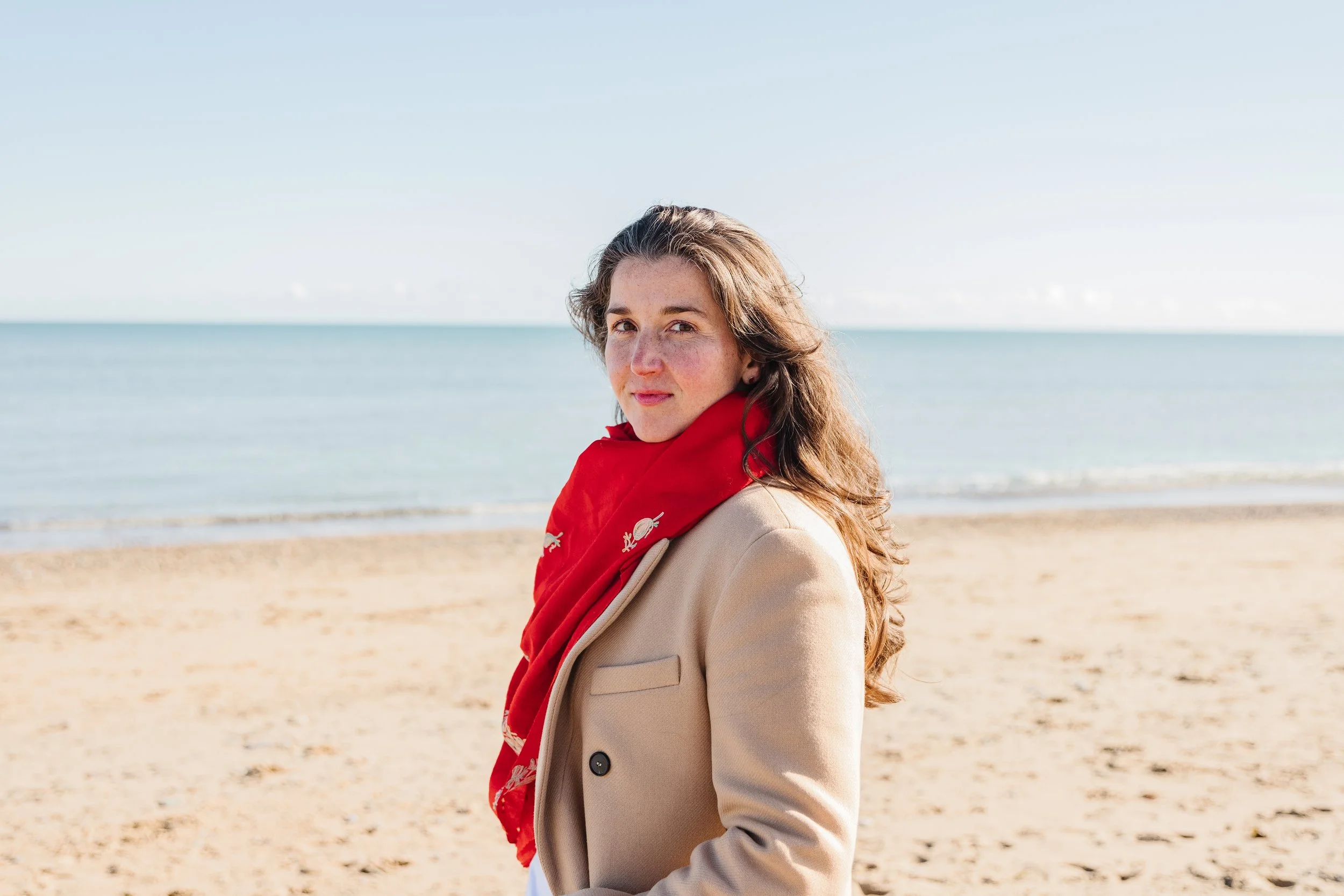 Jacqueline Kelly, perinatal psychotherapist and founder of Our Brave Hearts, standing on a beach wearing a red scarf and camel coat