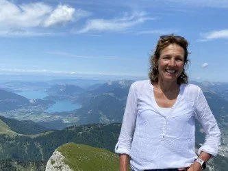 Vrouw staat op een bergtop met een glimlach, uitkijkend over bergen en meren onder een blauwe lucht.
