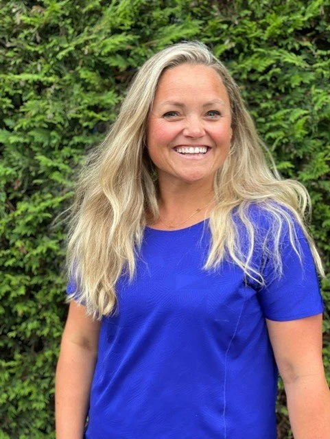 Vrouw met lang blond haar en een blauwe shirt, lachend, voor een groene struik