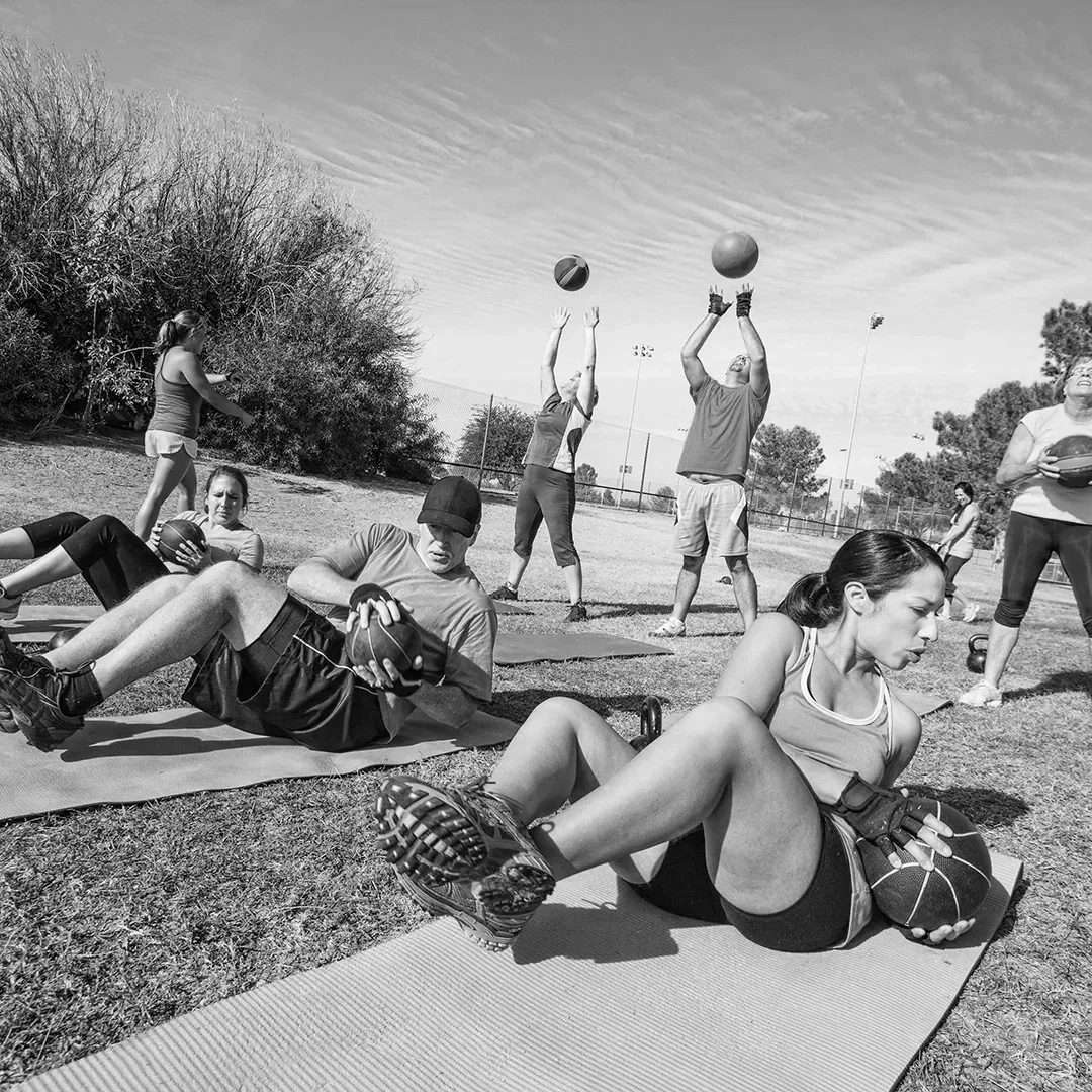 People exercising outdoors with medicine balls on mats, some throwing or holding balls, in a park with trees and a cloudy sky.