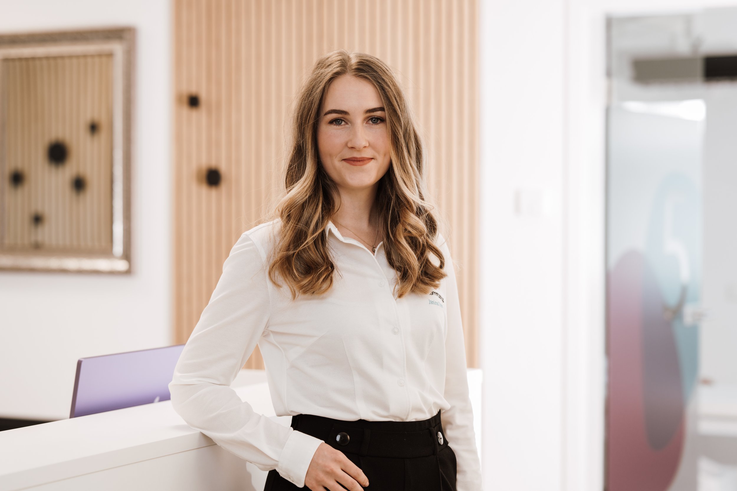Eine junge Frau mit langem, gewelltem braunem Haar steht an einem Empfangstresen in einem modernen Raum, trägt eine weiße Bluse und schwarze Hosen, lächelt leicht und blickt in die Kamera.