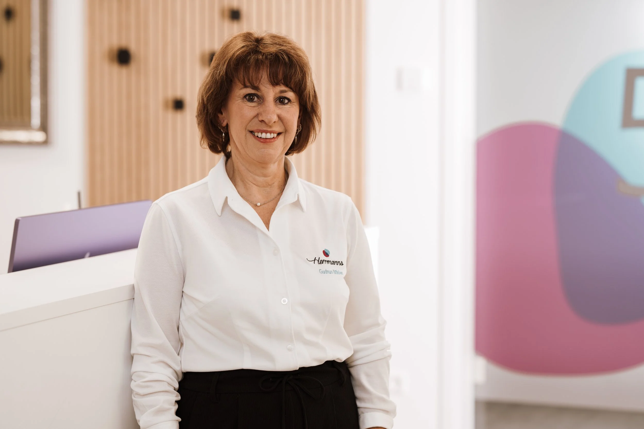 Eine lächelnde Frau in weißem Hemd steht im Empfangsbereich eines modernen Gebäudes, im Hintergrund eine bunte Wand und eine Arbeitsfläche.