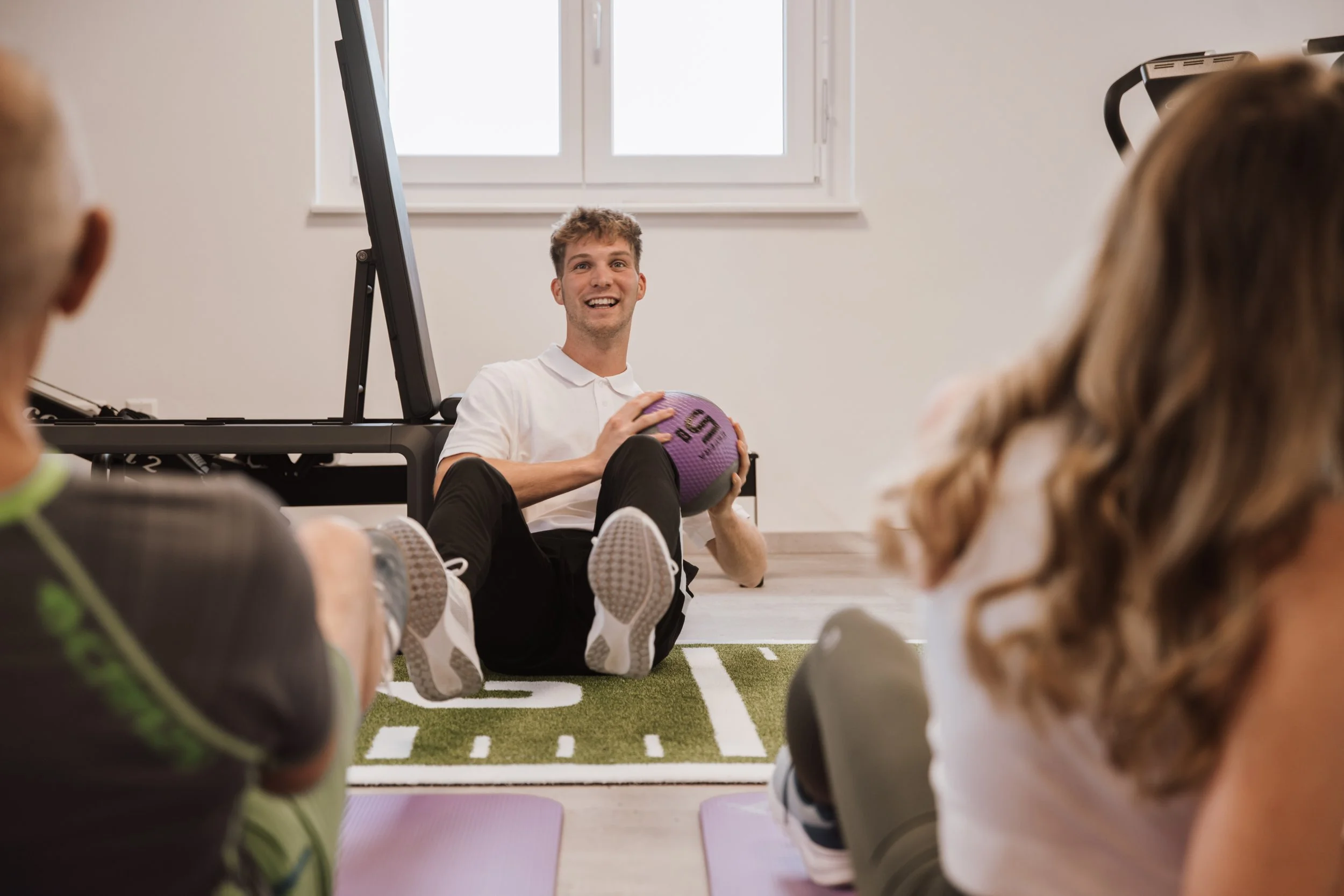 Ein junger Mann mit weißem T-Shirt sitzt auf dem Boden und hält einen purple Fitnessball in den Händen, während er vor einer Gruppe sitzt, die Yoga- oder Fitnessübungen auf Matten macht. Das Zimmer ist hell mit einem Fenster im Hintergrund.