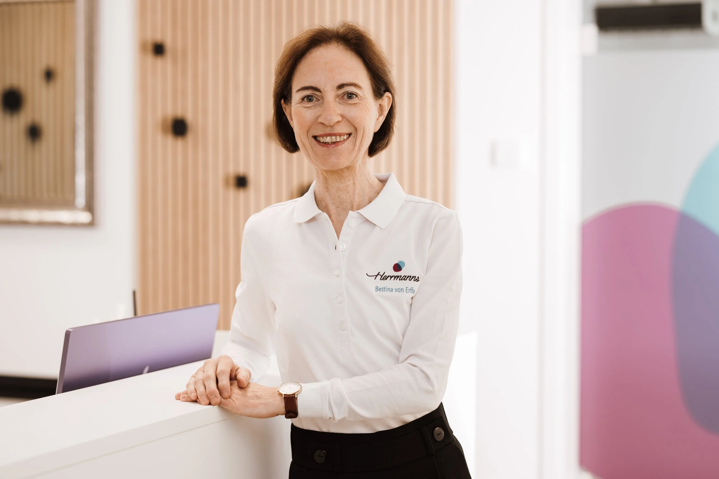 Eine lächelnde Frau steht an einem Empfangstisch in einem modernen Raum, trägt ein weißes Hemd mit einem Logo, das 'Herrmanns' heißt, und sieht zur Kamera.