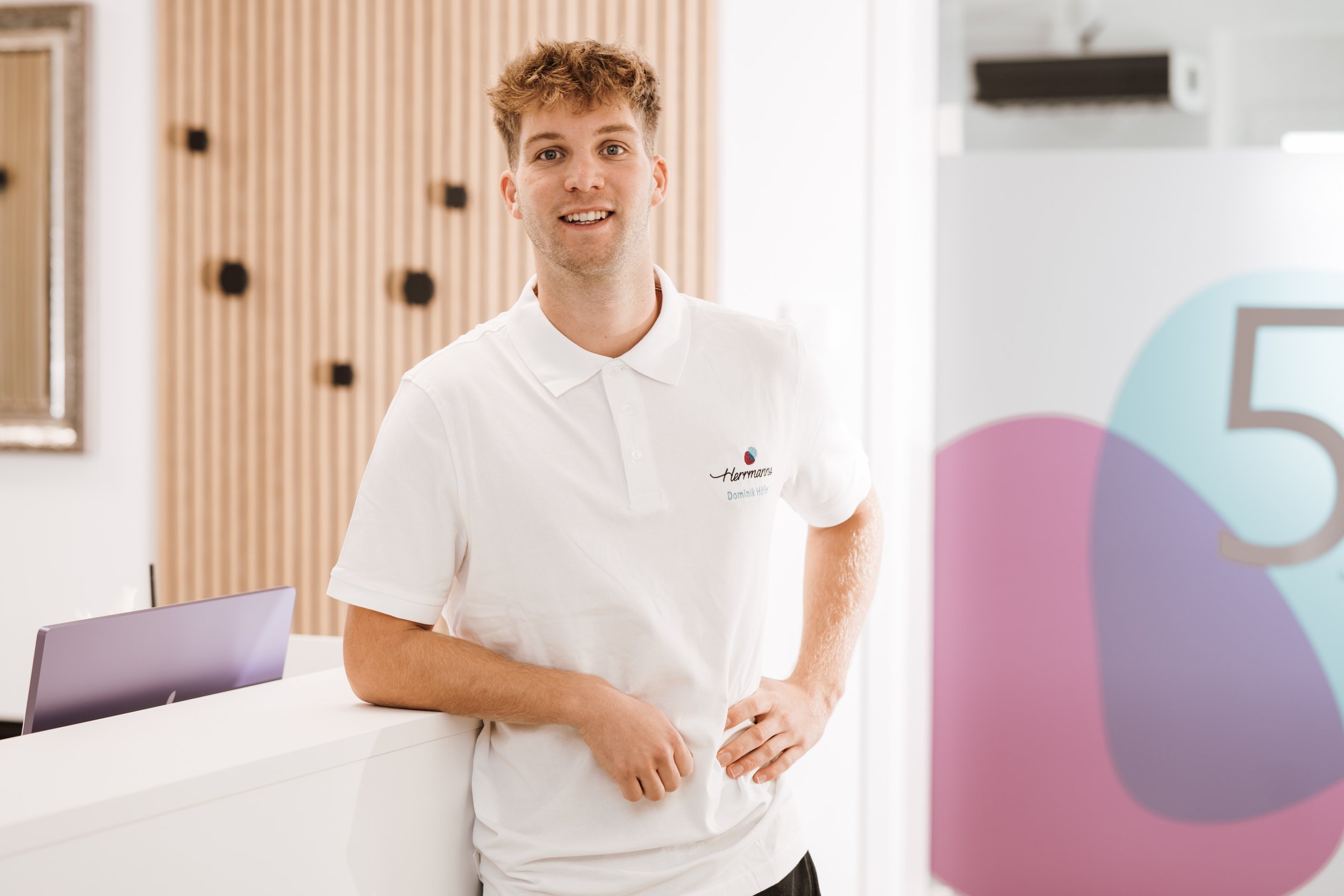 Ein junger Mann in einem weißen Polo-Shirt steht in einem modernen Empfangsbereich.
