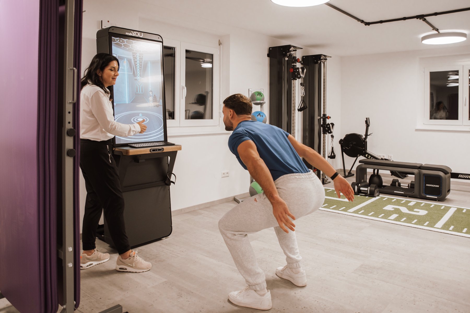 Ein Mann trainiert im Fitnessstudio mit einem Personal Trainer, während er eine virtuelle Übung auf einem Bildschirm ausführt.
