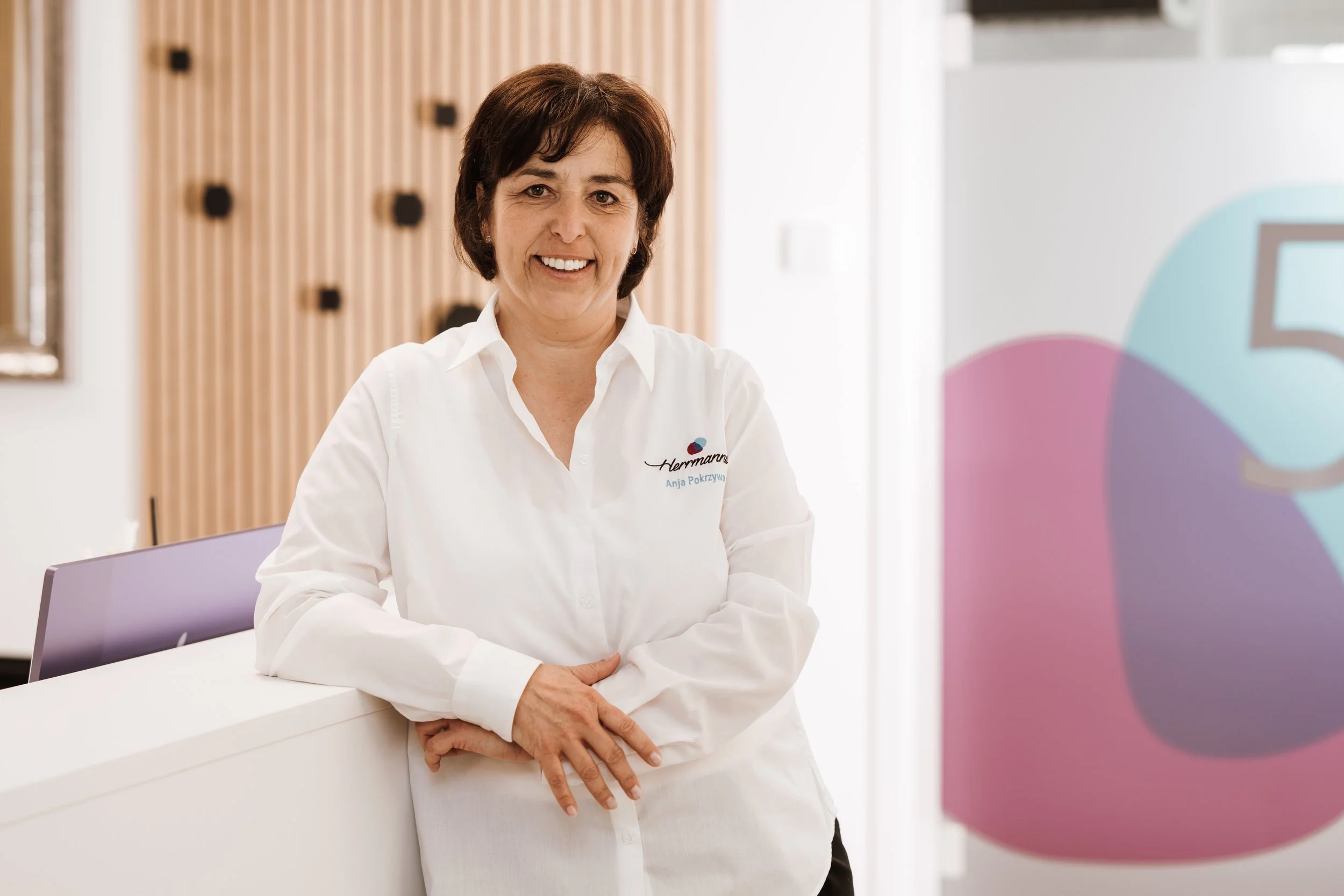 Eine Frau mit dunklen Haaren, trägt ein weißes Hemd mit einem Logo und Namen, steht in einem modernen Raum mit Holzdekor und lächelt.