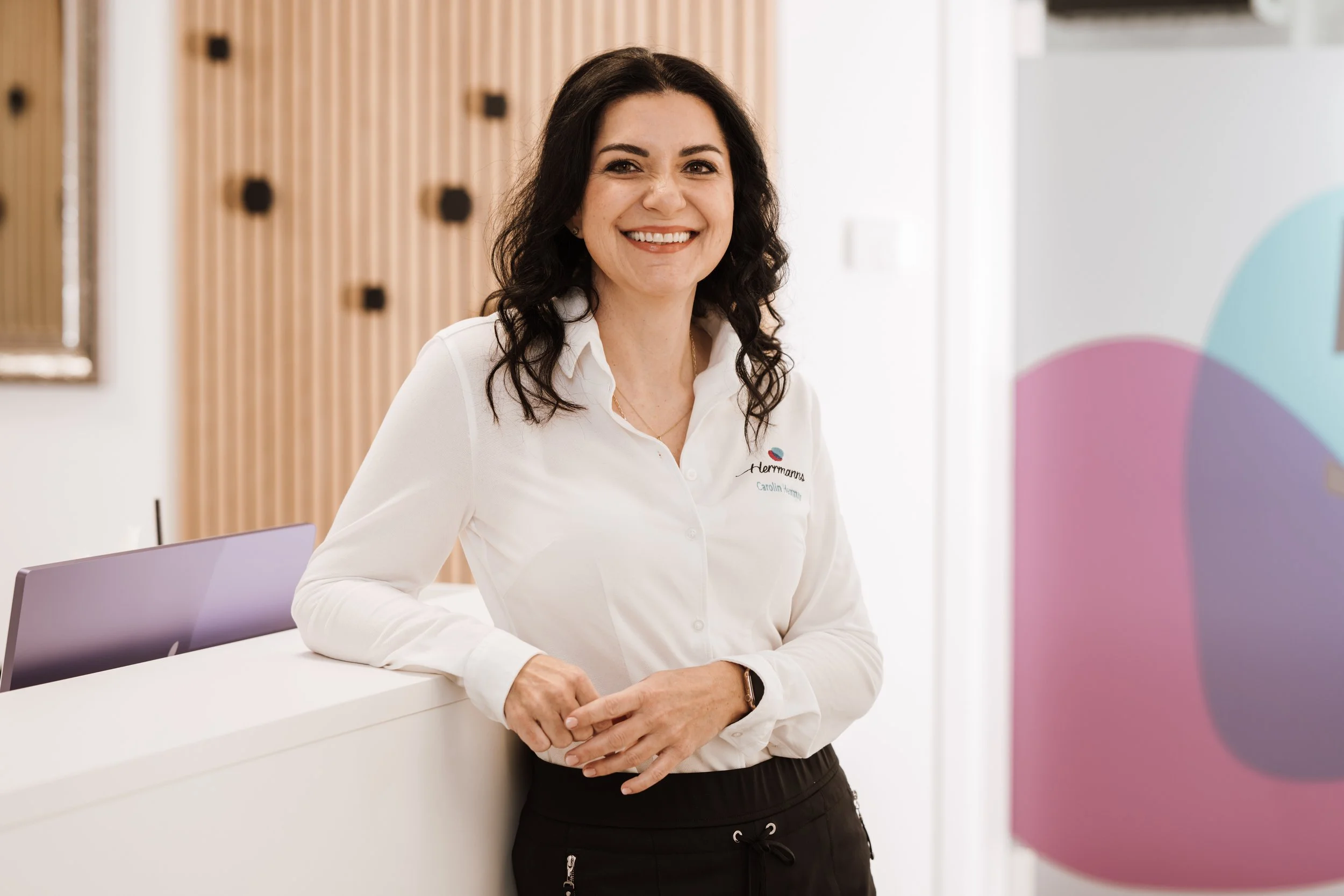 Lächelnde Frau mit dunklem Haar in weißer Bluse steht an einem Empfangstresen in einem modernen, hellen Raum.