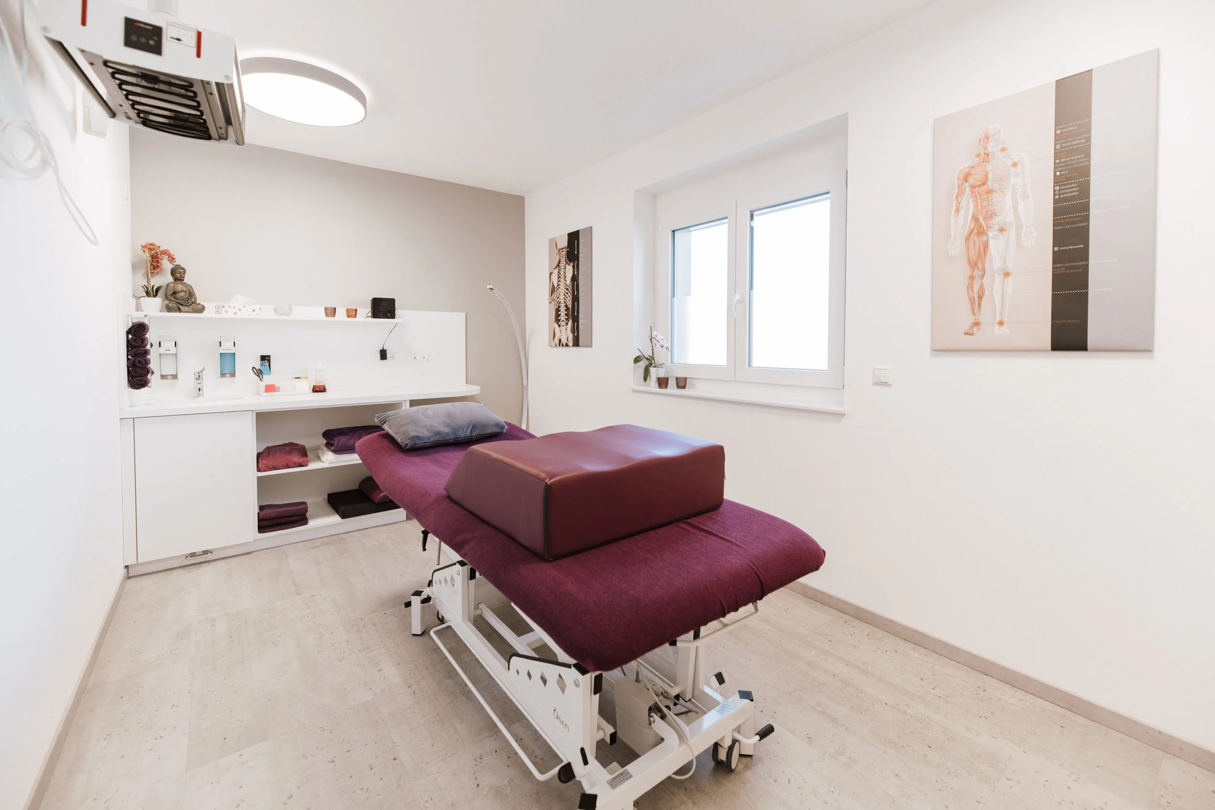 Behandlungsraum in einer medizinischen Praxis mit Behandlungsliege, medizinischen Utensilien und anatomischen Postern an den Wänden.