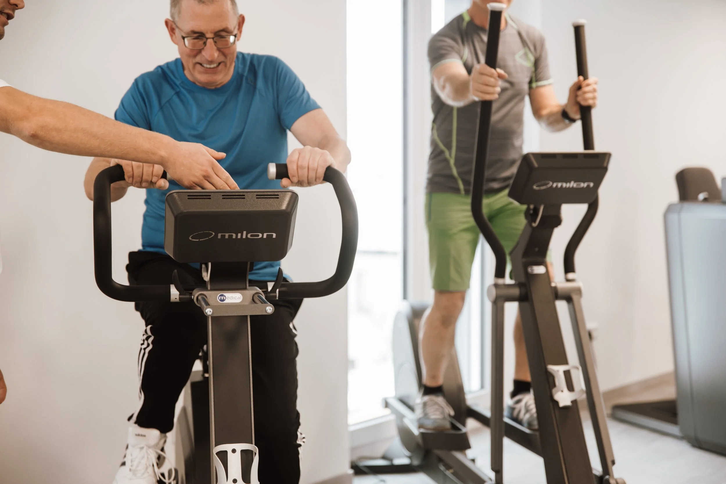 Zwei ältere Männer trainieren mit Fitnessgeräten im Fitnessstudio. Der Mann auf der linken benutzt ein stationäres Fahrrad und lächelt, während ein Trainer ihm assistiert. Der Mann auf der rechten benutzt ein Steppgerät und trägt graues T-Shirt und g