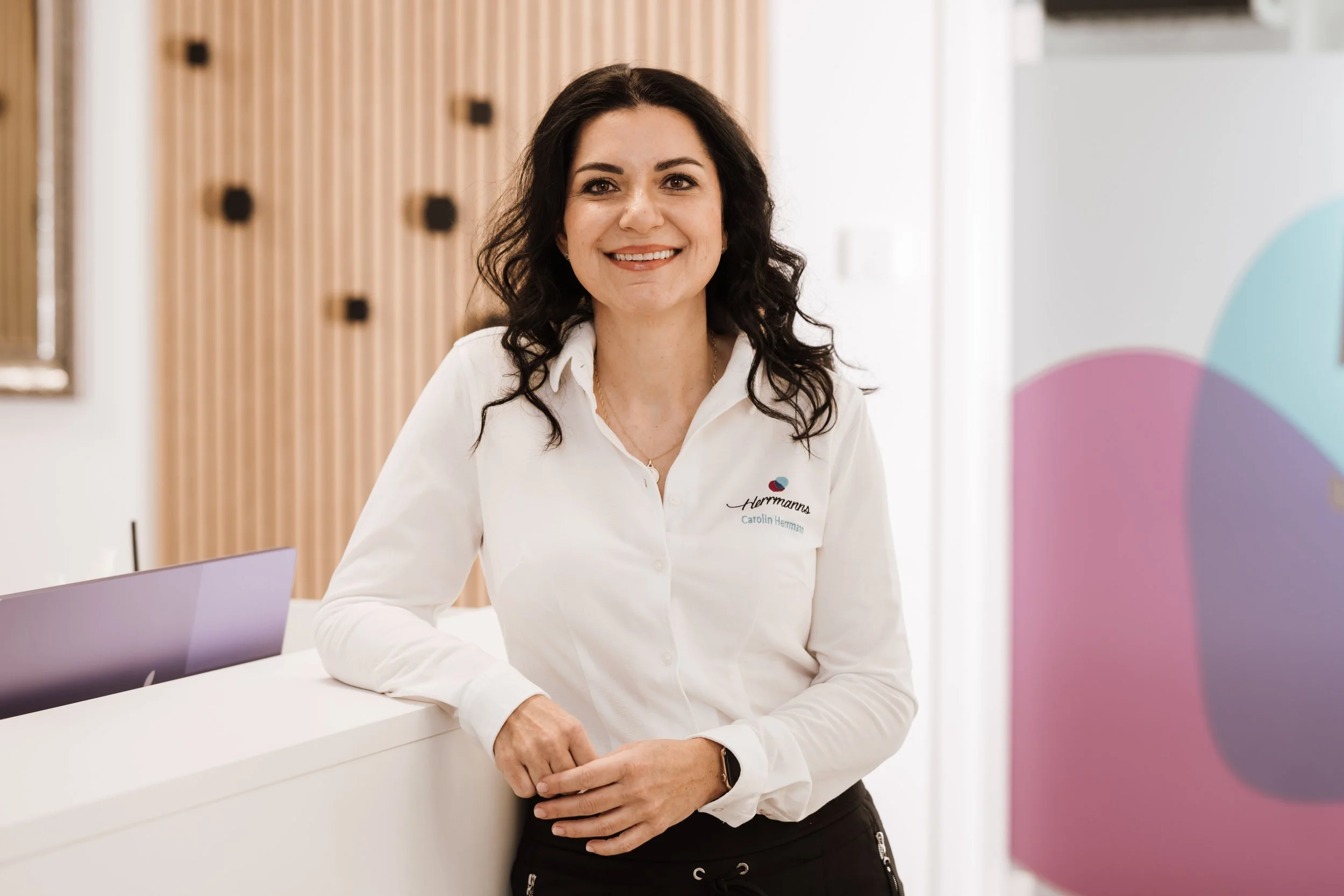 Eine lächelnde Frau mit dunklen, lockigen Haaren trägt eine weiße Bluse mit Logo und Name an der Rezeption eines modernen Büros.