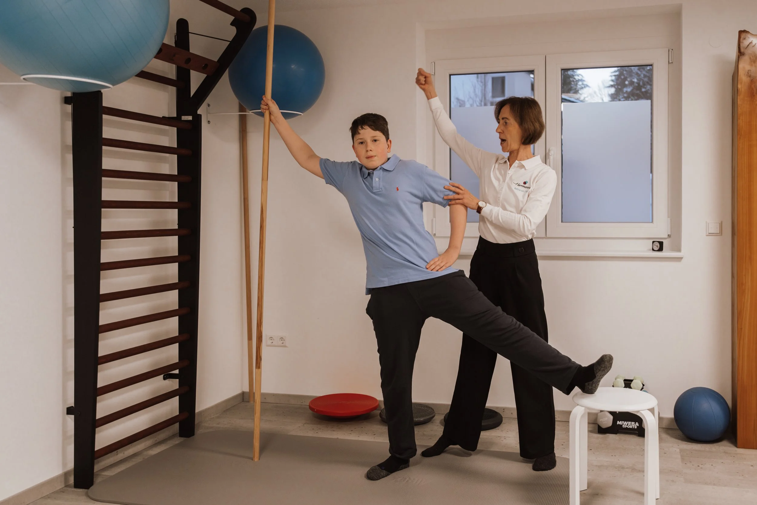 Ein Therapeut, der einem Jungen bei einer Physiotherapie-Übung hilft, während der Junge eine Pose mit einem Bein hebt und den Arm mit einem Stock nach oben streckt, in einem Therapieraum mit Fitnessgeräten und Bällen.
