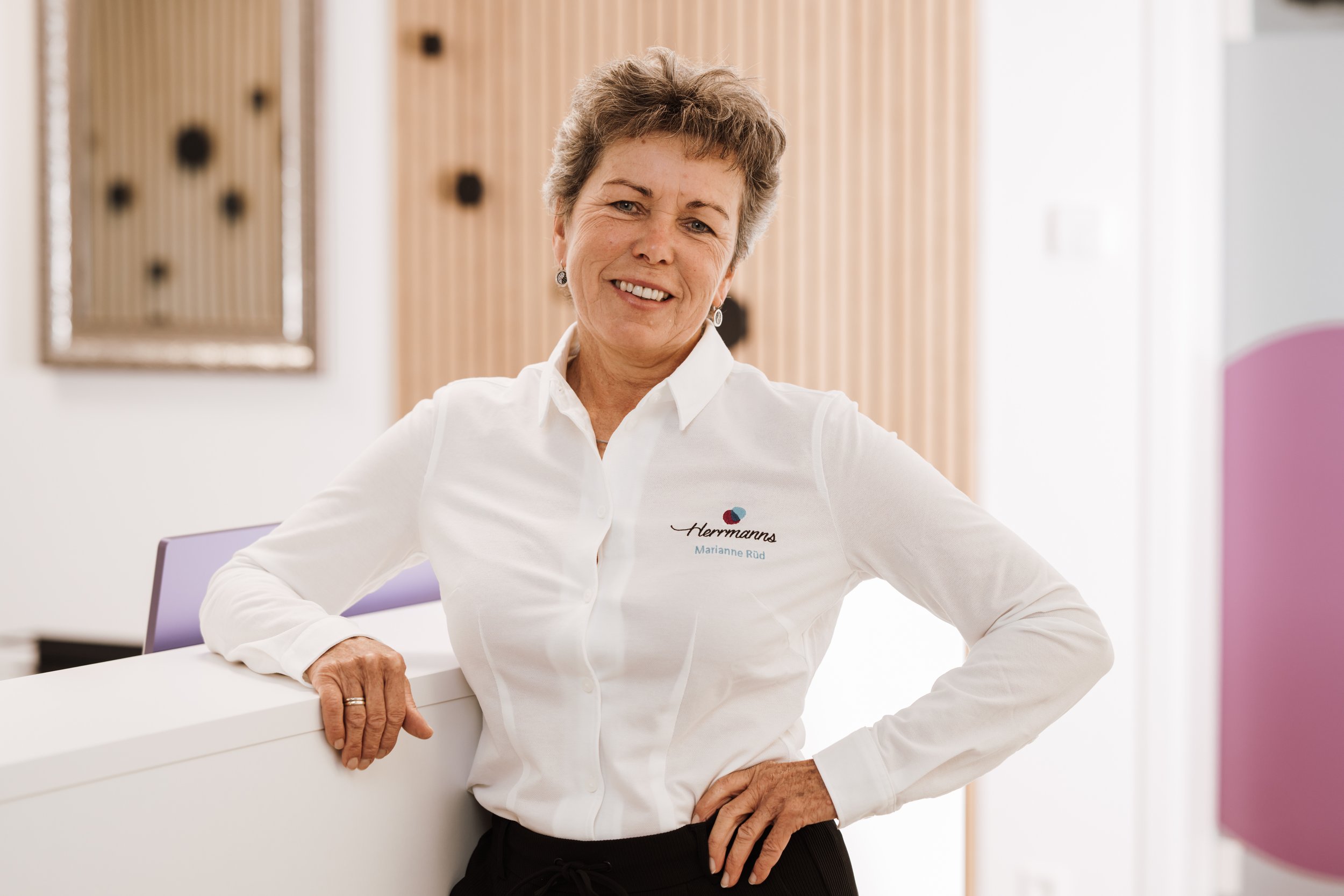 Eine lächelnde ältere Frau in einem weißen Hemd steht in einem modernen Büro, lehnt an einem weißen Tresen, mit einer Rückenlehne in Lila, im Hintergrund.