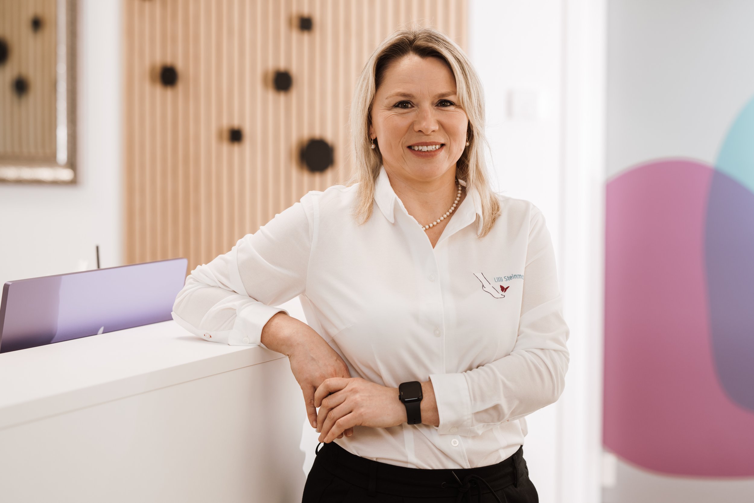 Eine lächelnde Frau steht an einer Rezeption, trägt eine weiße Hemd mit Logo, eine Armbanduhr, Schmuck und hat blonde Haare.