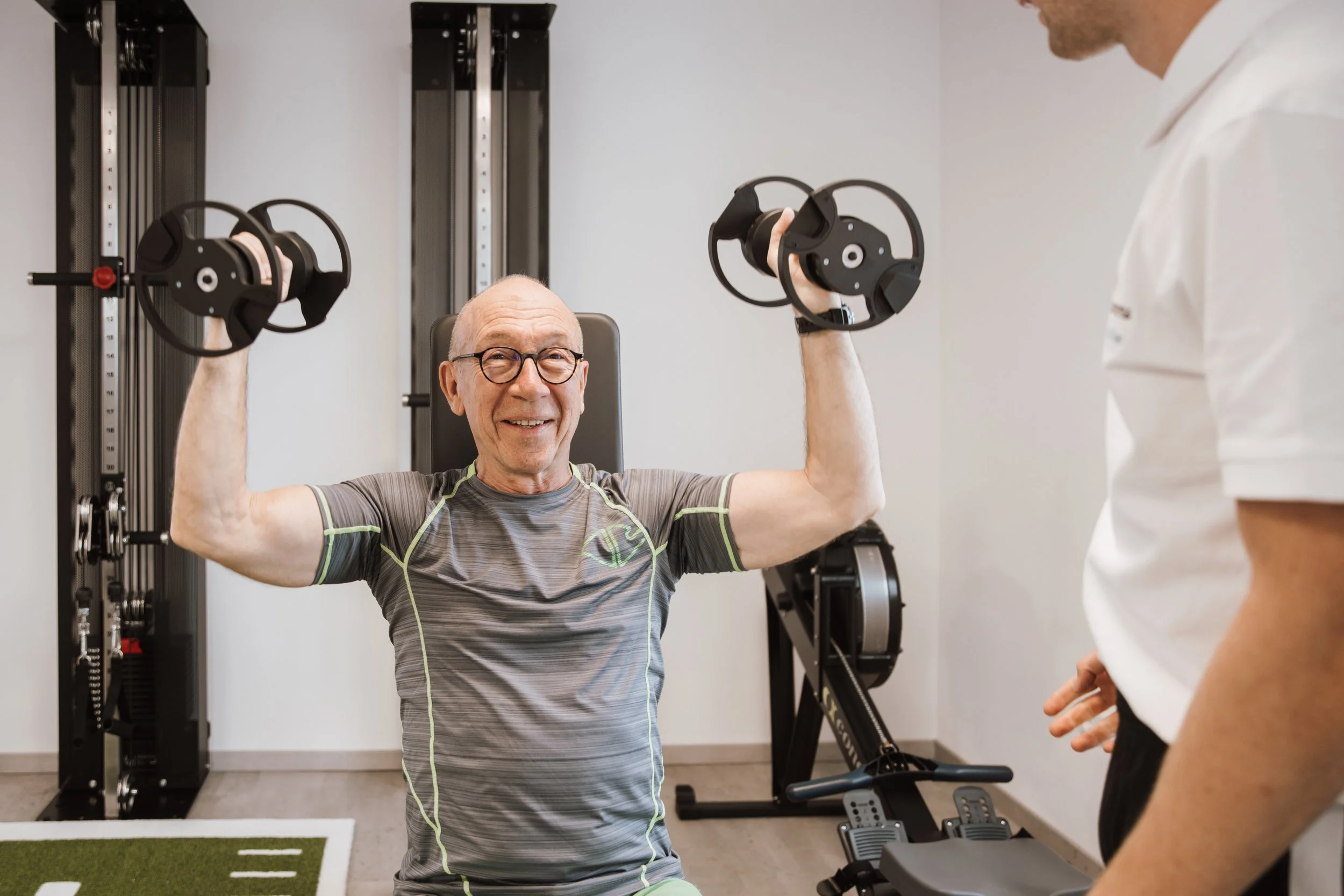Ein älterer Mann beim Krafttraining im Fitnessstudio, der mit Kurzhanteln übt, während ein Trainer ihm zuschaut.
