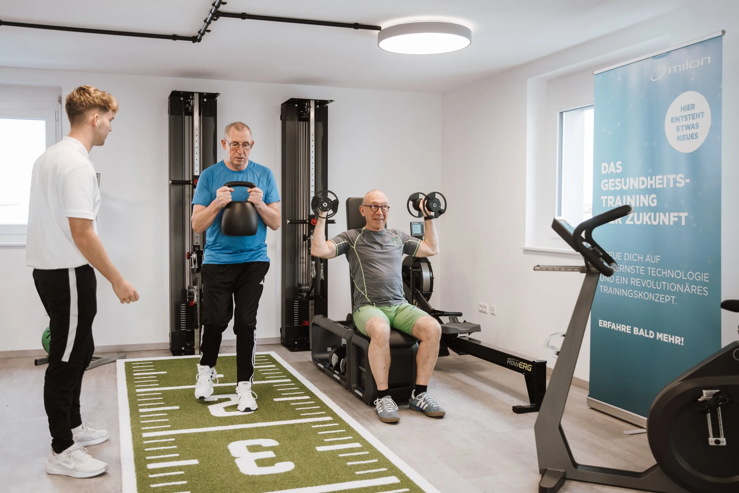 Ein Mann trainiert auf einem Fitnessgerät, während zwei weitere Personen ihn beobachten, in einem Fitnessraum mit Geräten und einer blauen Werbetafel.