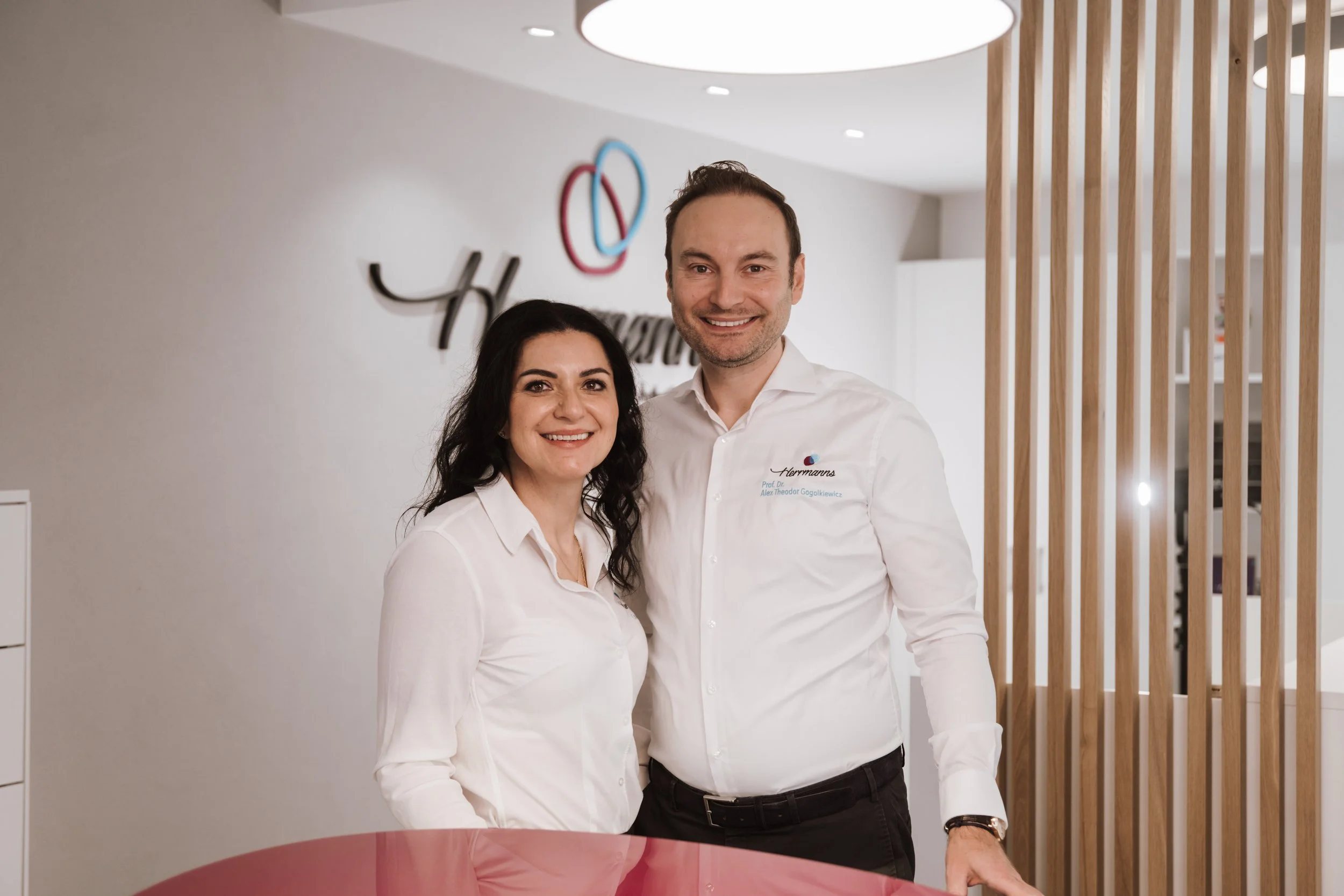 Zwei Personen, ein Mann und eine Frau, stehen hinter einem Pinken Empfangstresen in einem hellen Raum, lächeln und schauen in die Kamera. Der Mann trägt ein weißes Hemd mit einem Logo und Text, die Frau trägt ebenfalls ein weißes Hemd. Hintergrund zeigt eine weiße Wand mit einem Firmenlogo und Holzlamellen als Raumdekoration.