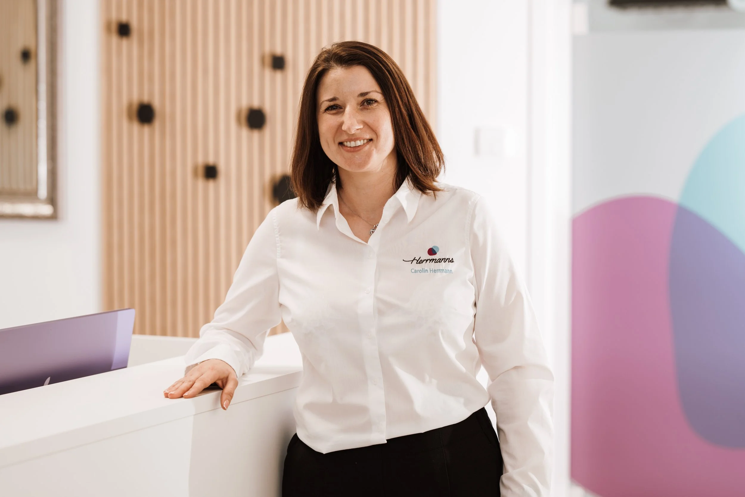 Eine lächelnde Frau in einer weißen Bluse mit Logo an der Rezeption eines modern eingerichteten Büros.