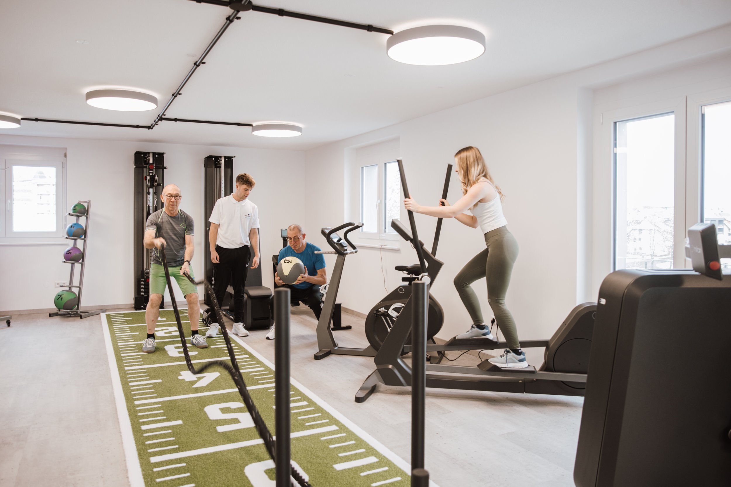 Menschen trainieren in einem Fitnessstudio, darunter eine Frau auf einem Laufband, ein Mann mit einer Gewichtsscheibe, ein Mann mit einem Seil und ein Trainer, der einen Medizinball hält.